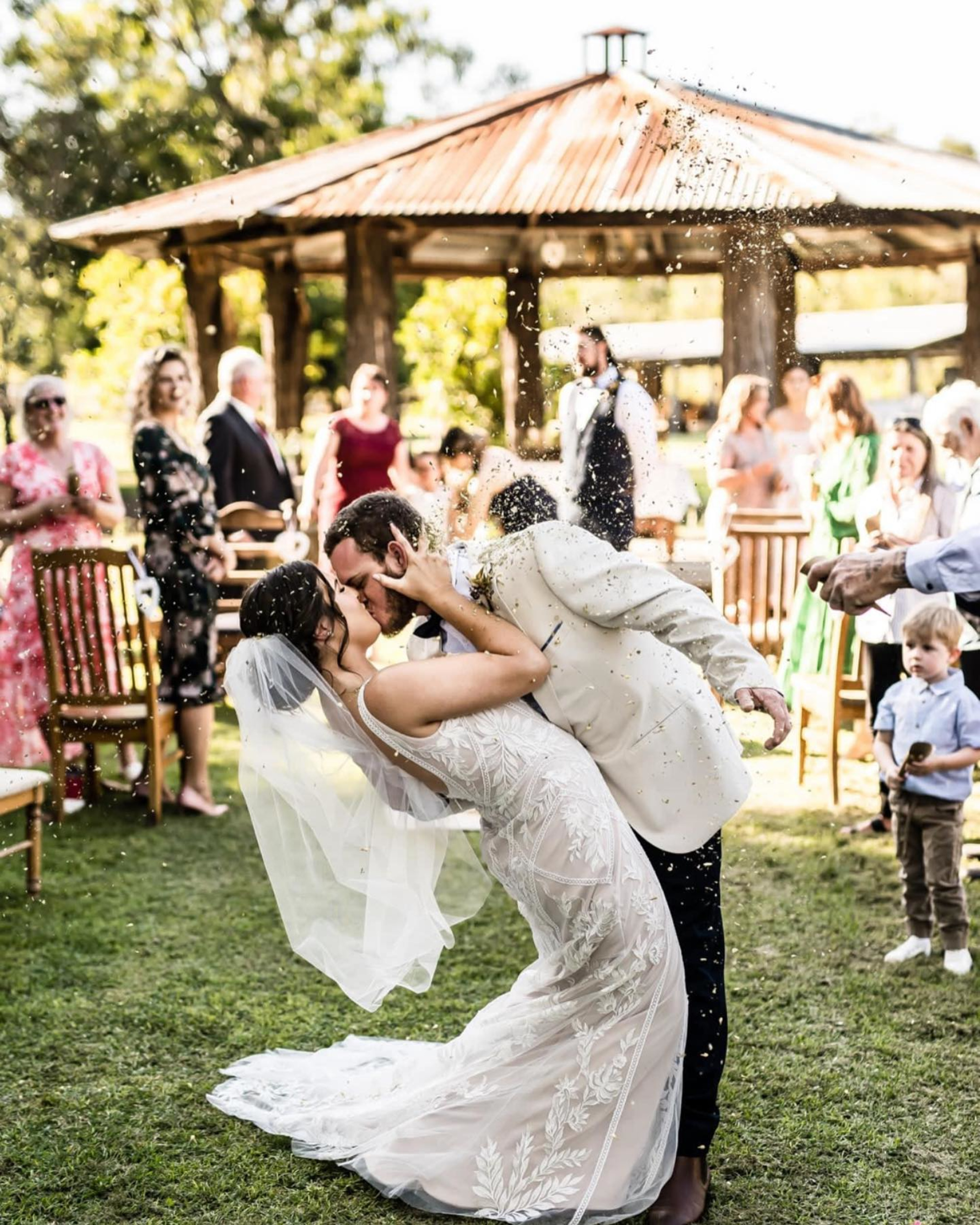 Newlywed couple shares a dip kiss outdoors as guests toss confetti at a rustic garden wedding ceremony.