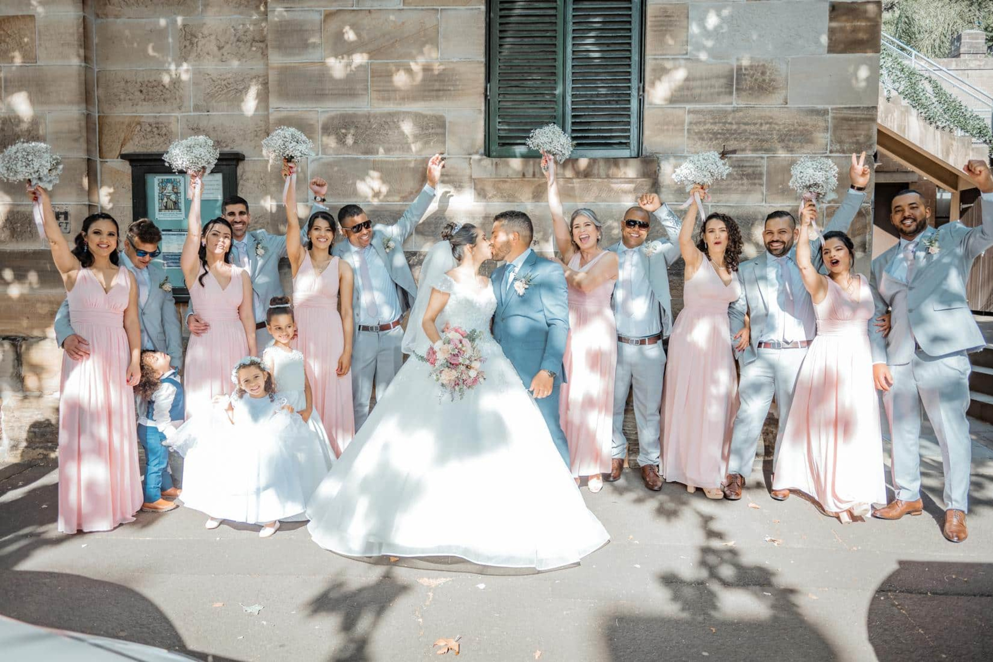 Joyful wedding party in pastel pink and blue outfits cheering around the kissing bride and groom outdoors.