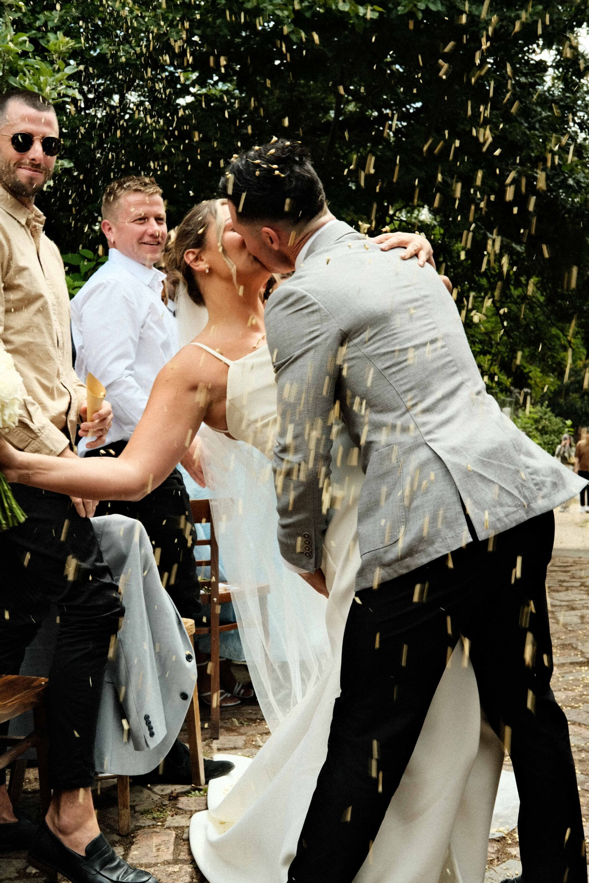 Bride and groom share a kiss during an outdoor confetti toss surrounded by guests.