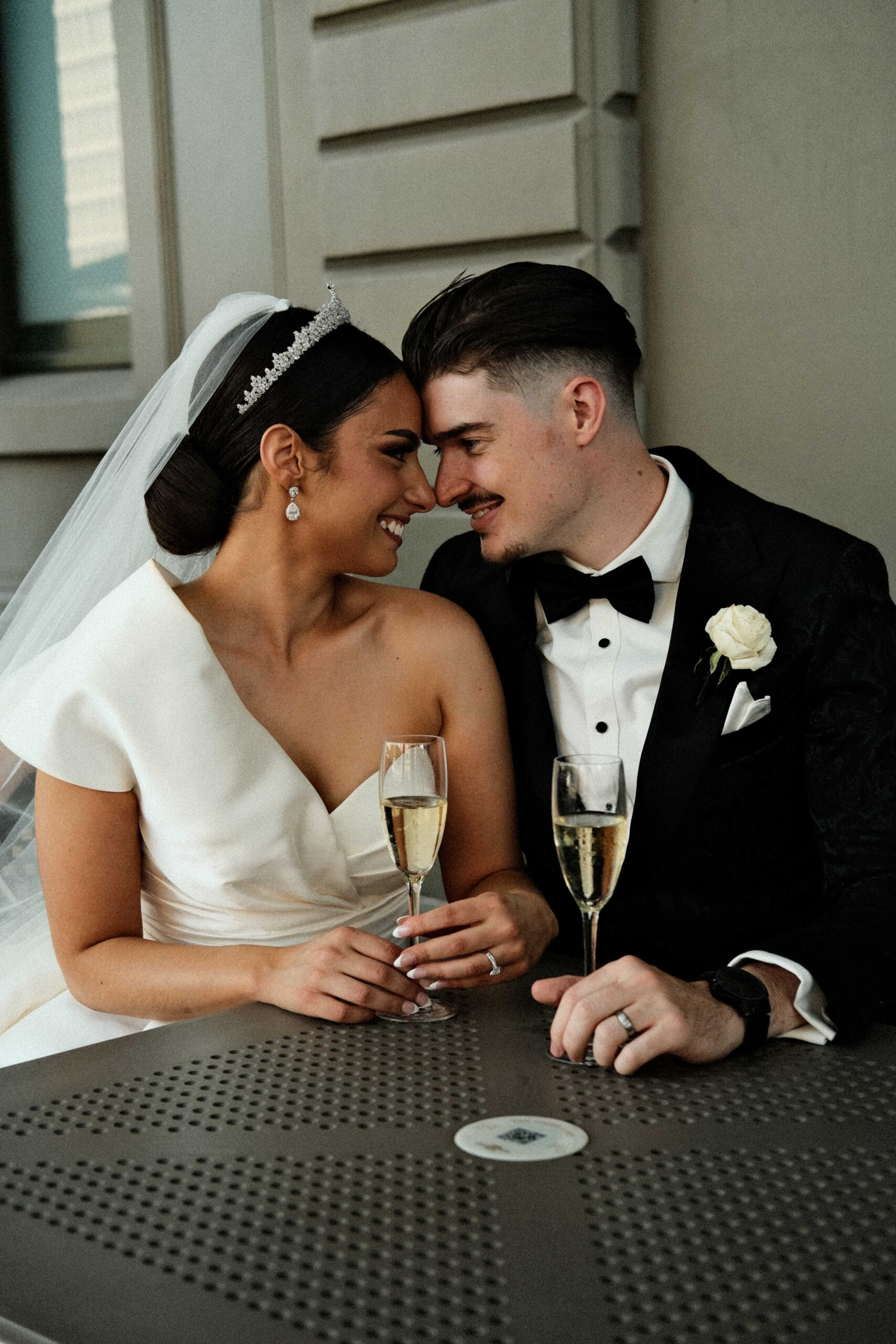 Smiling bride and groom in formal attire touch foreheads while holding champagne glasses at their wedding.
