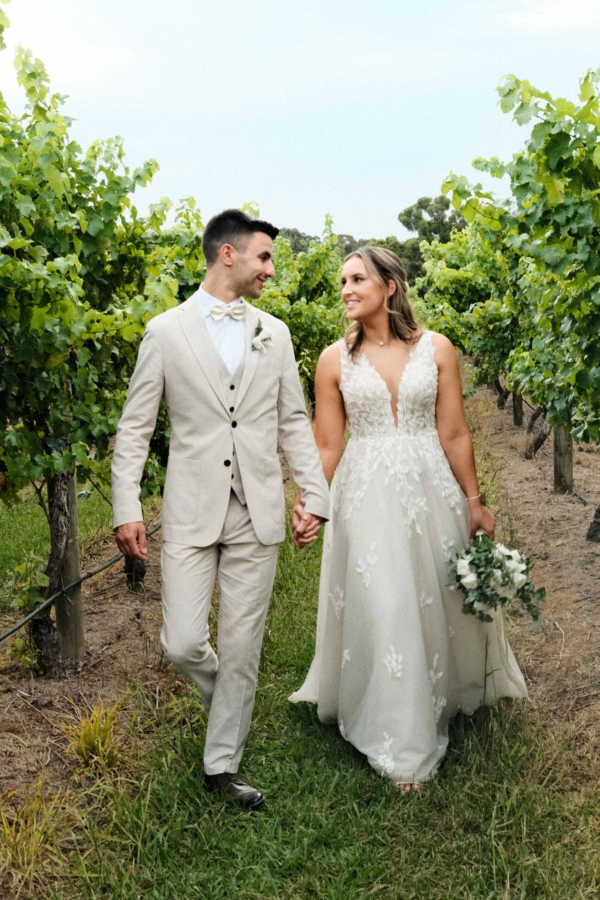 Bride and groom walk hand in hand through a lush vineyard on their wedding day.