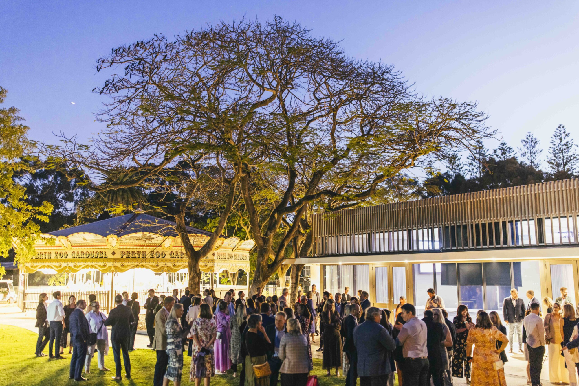 Evening outdoor wedding reception with guests mingling near a lit carousel and modern venue building under a large tree.