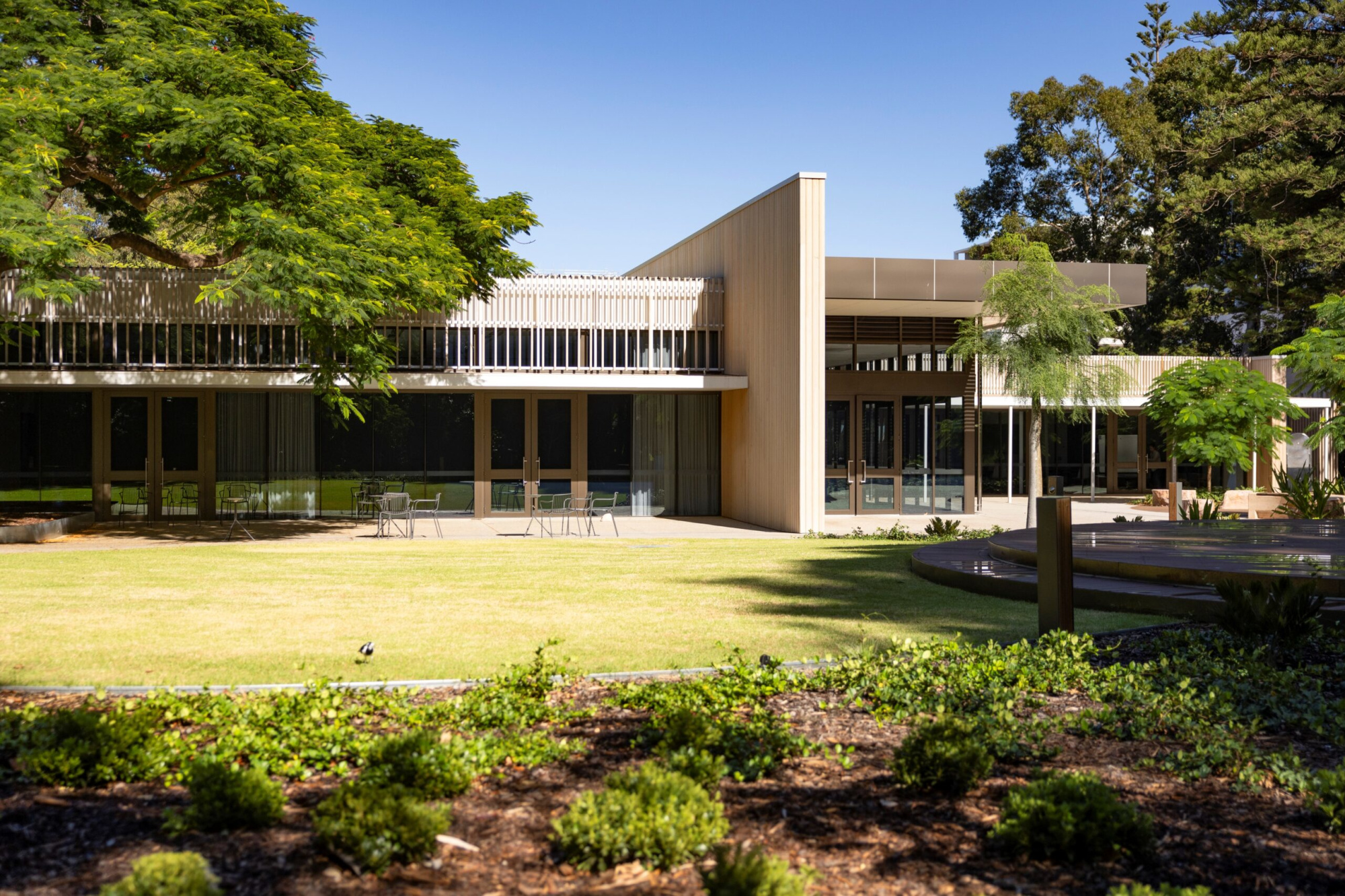 Modern event building with large windows overlooking a sunny lawn and landscaped garden, ideal for outdoor weddings.