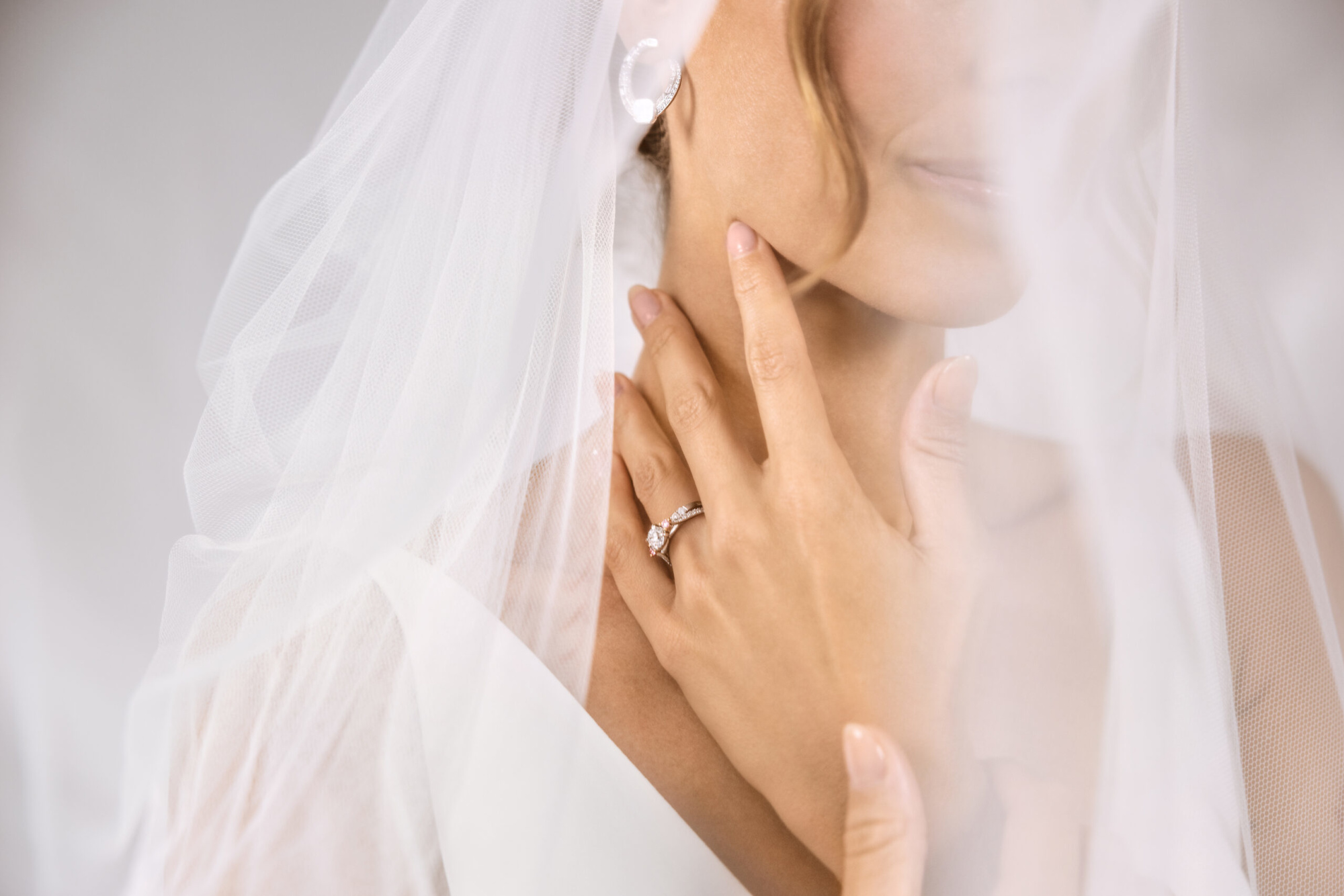 Close-up of a bride touching her neck, showcasing her engagement ring through a soft white veil.