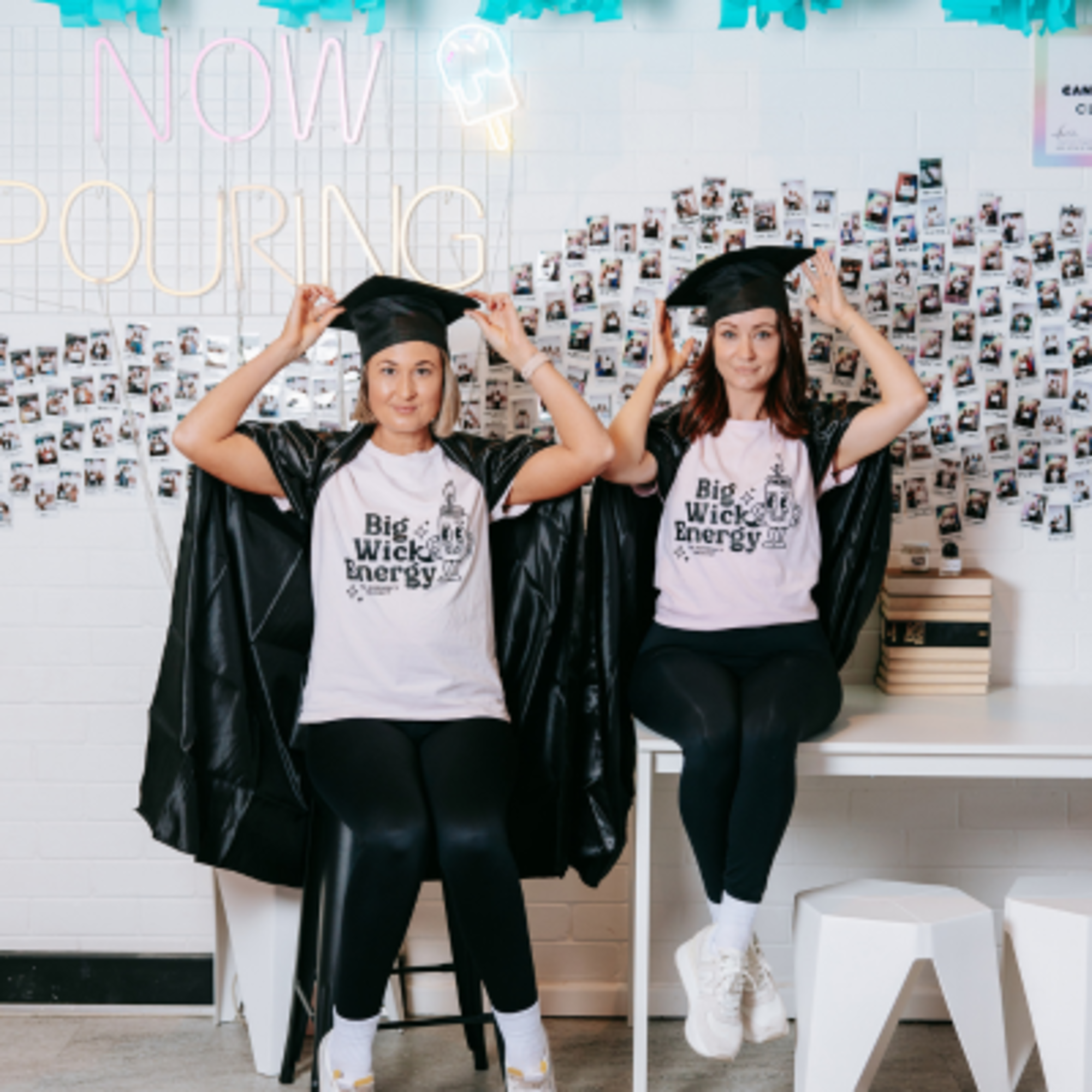 Two women in matching shirts and graduation caps pose playfully in front of a photo wall and neon sign.