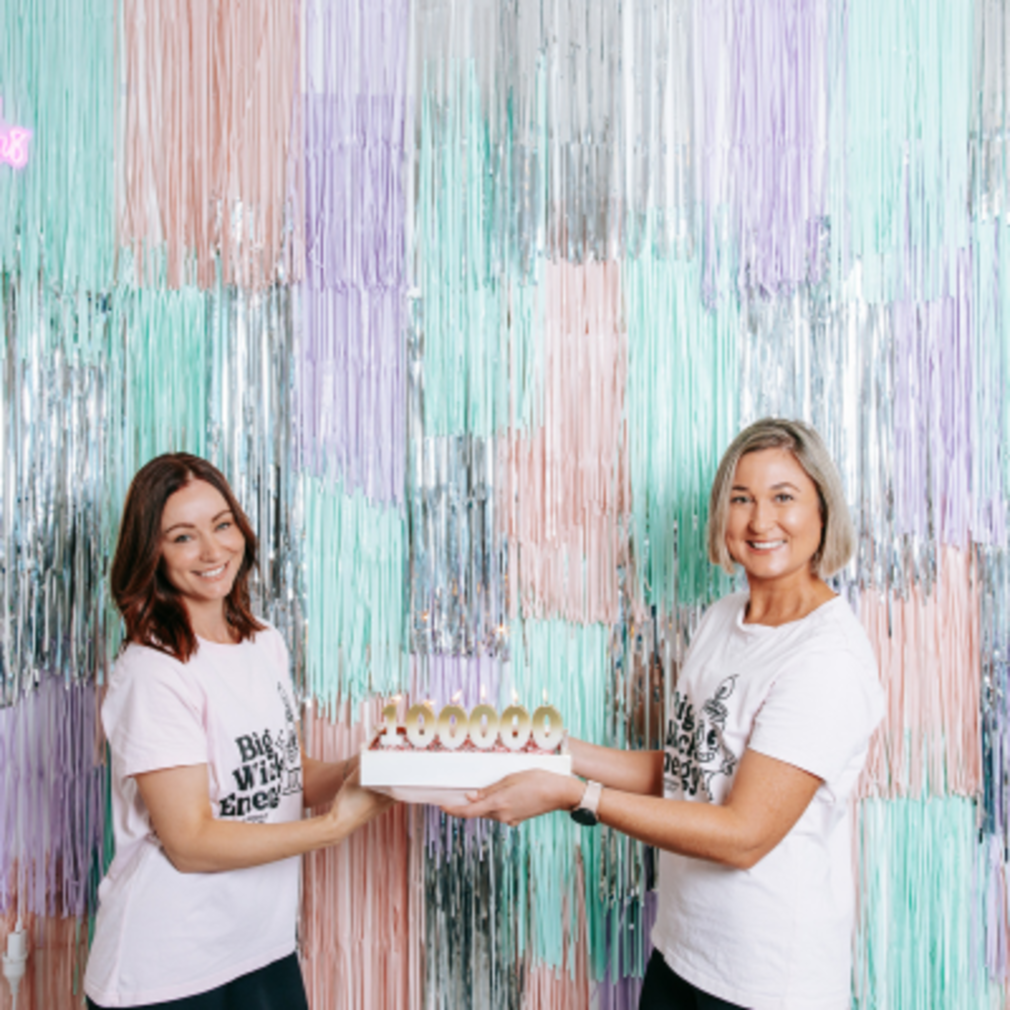 Two women hold a cake in front of a colorful pastel and silver fringe backdrop.