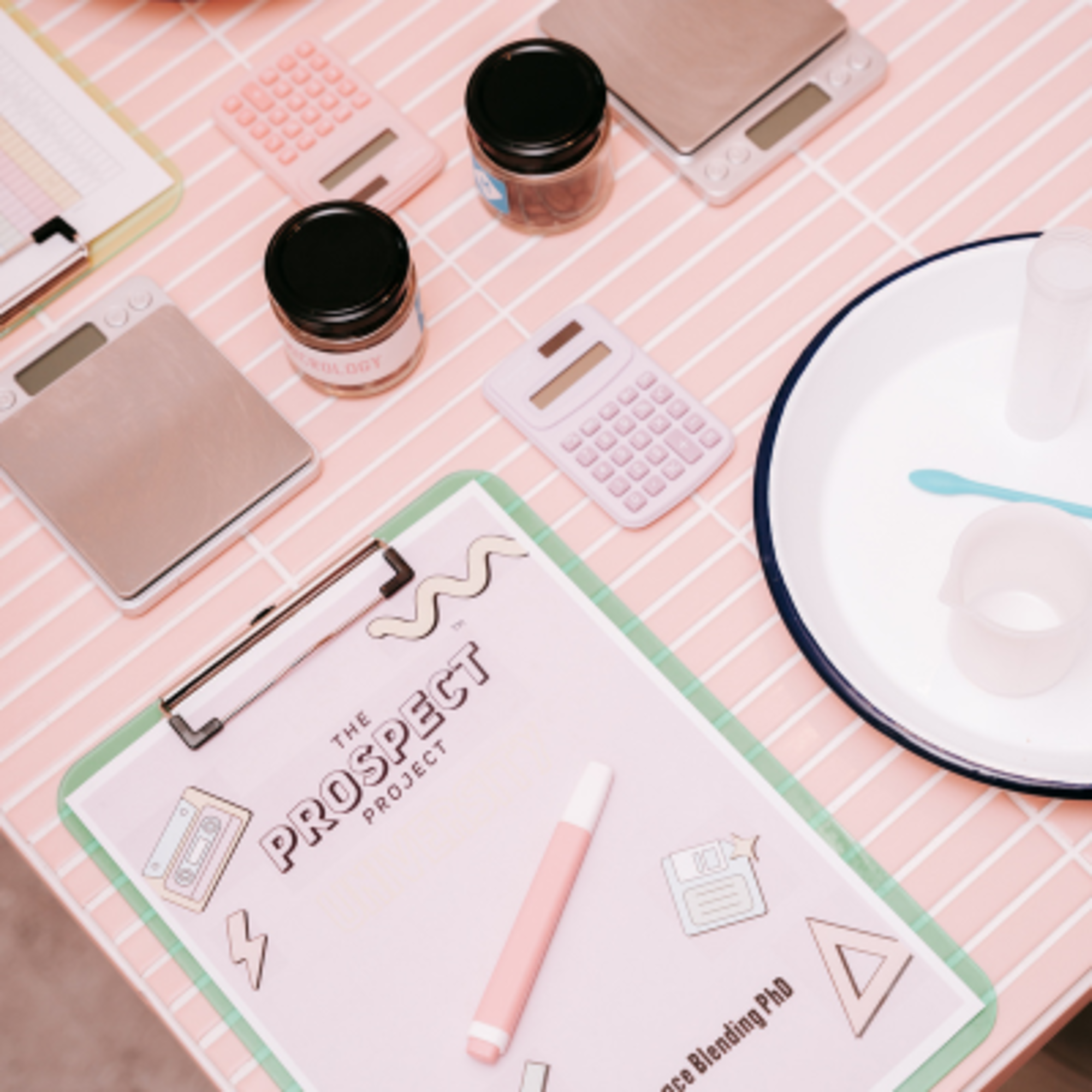 Styled flat lay of a wedding planning workspace with clipboard, calculators, scales, and jars on a pink tiled surface.