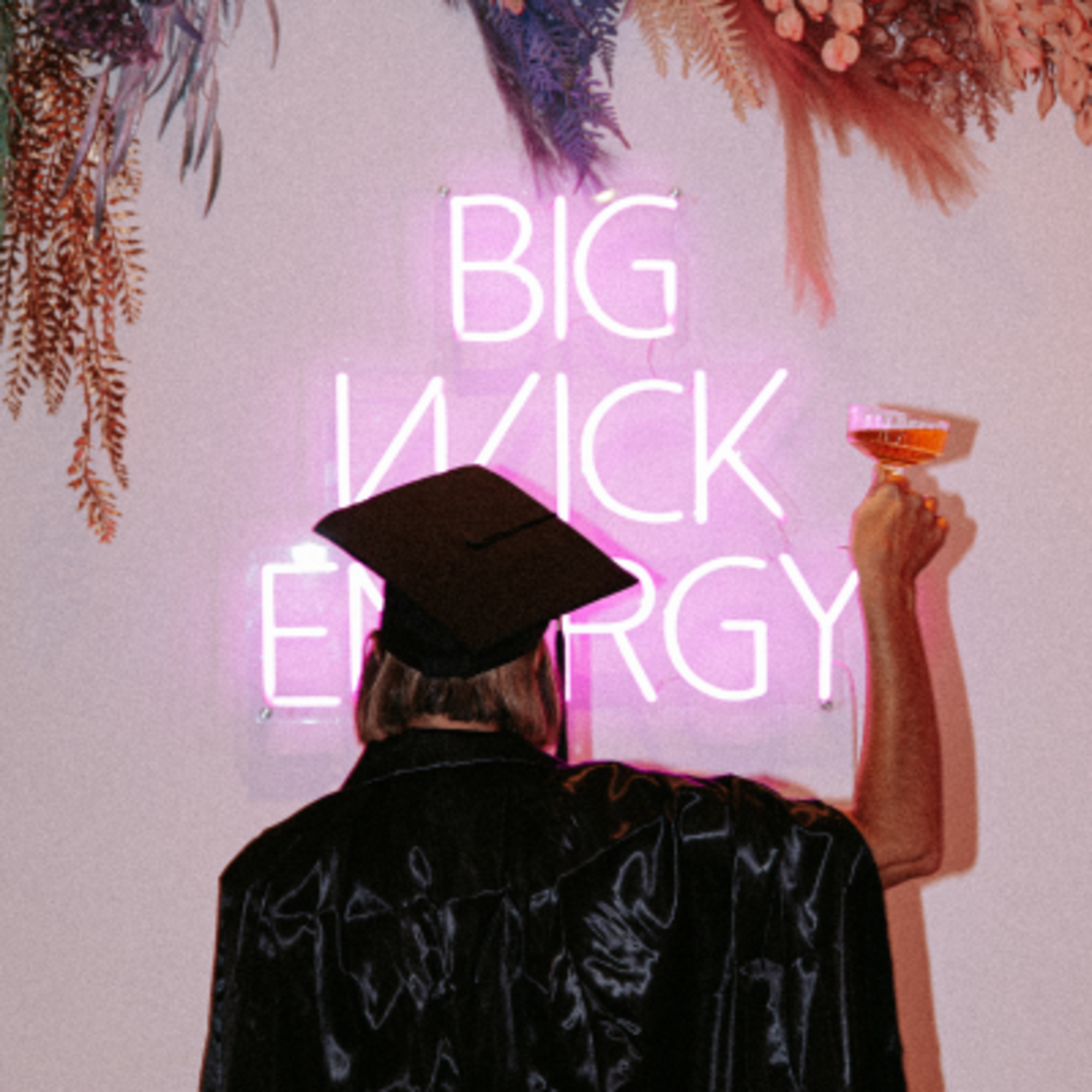 Person in a cap and gown raises a cocktail in front of a pink neon sign and hanging florals.
