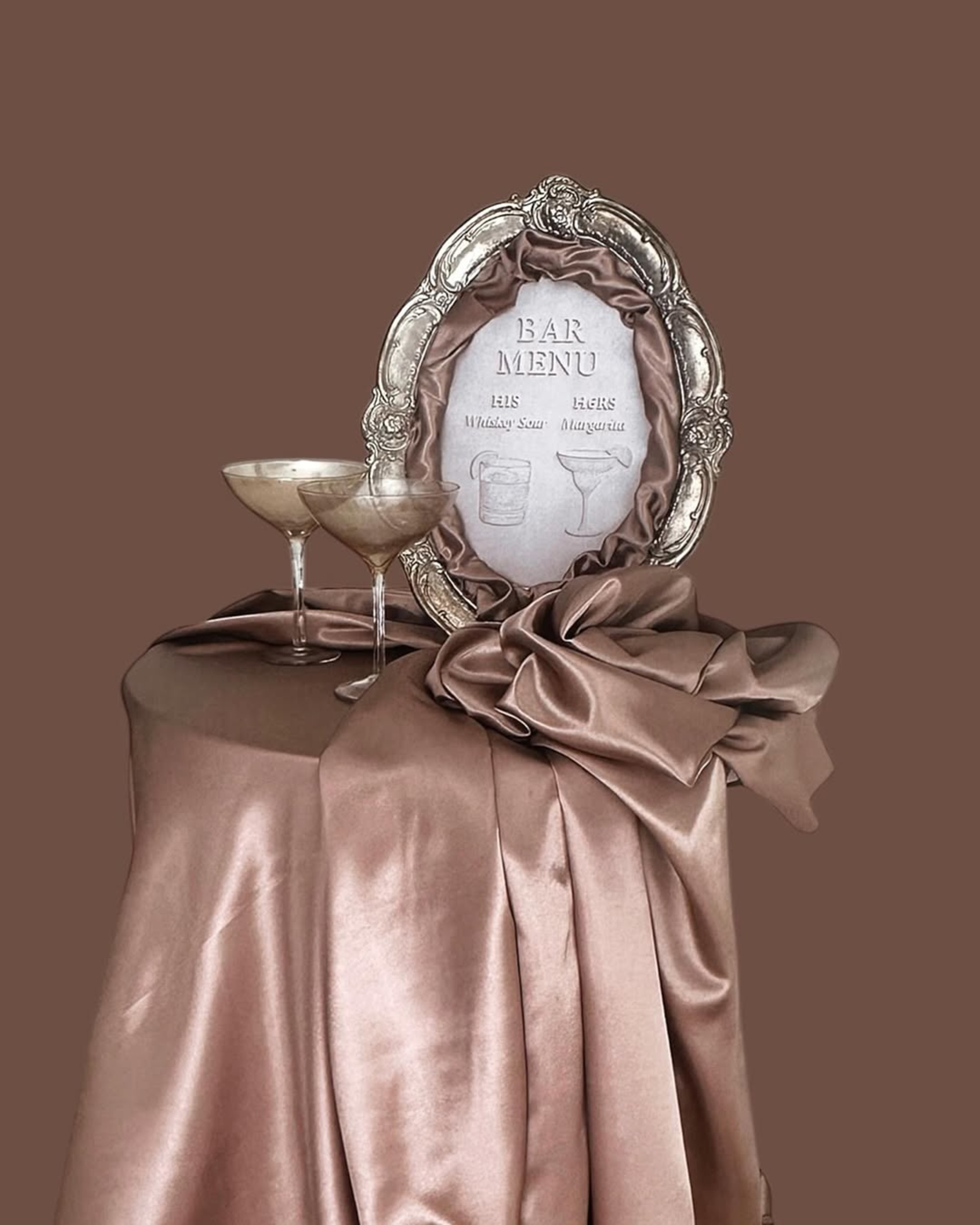 Elegant wedding bar menu in an ornate frame with two cocktail glasses on a satin-draped table.