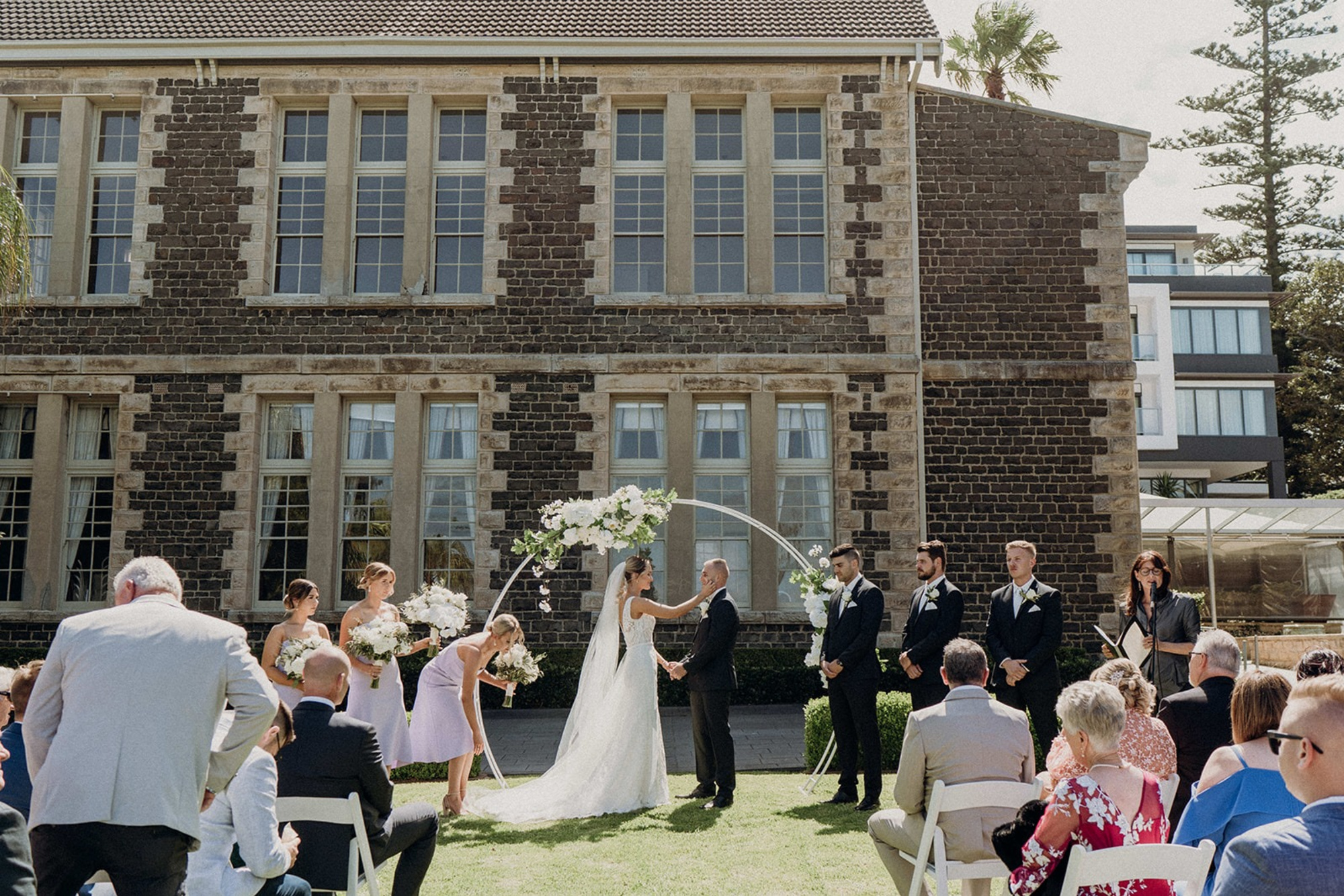 Outdoor wedding ceremony in front of a historic stone building with a floral arch and seated guests.