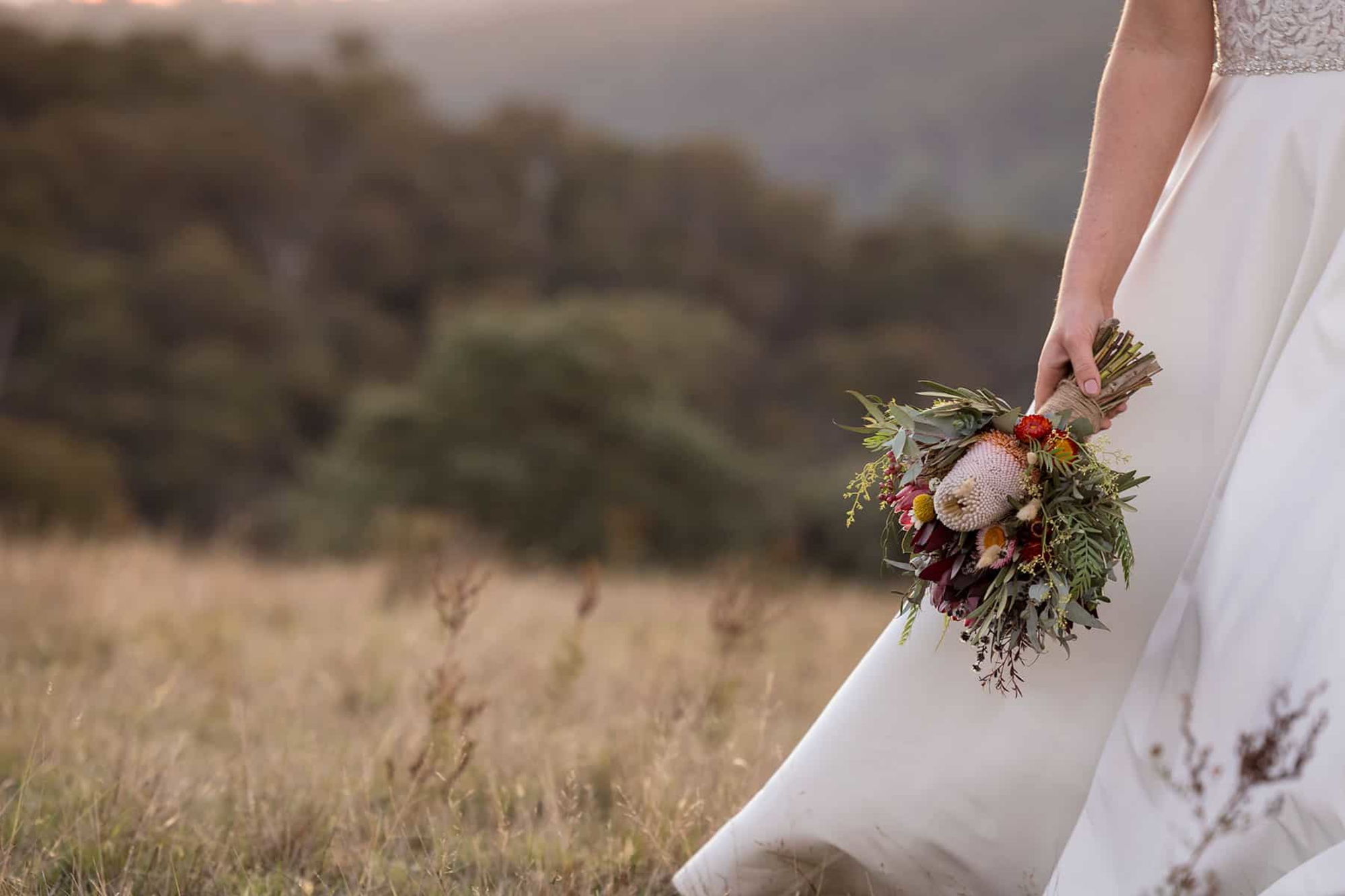 Bride holding a rustic wildflower bouquet while standing in a grassy field at sunset.