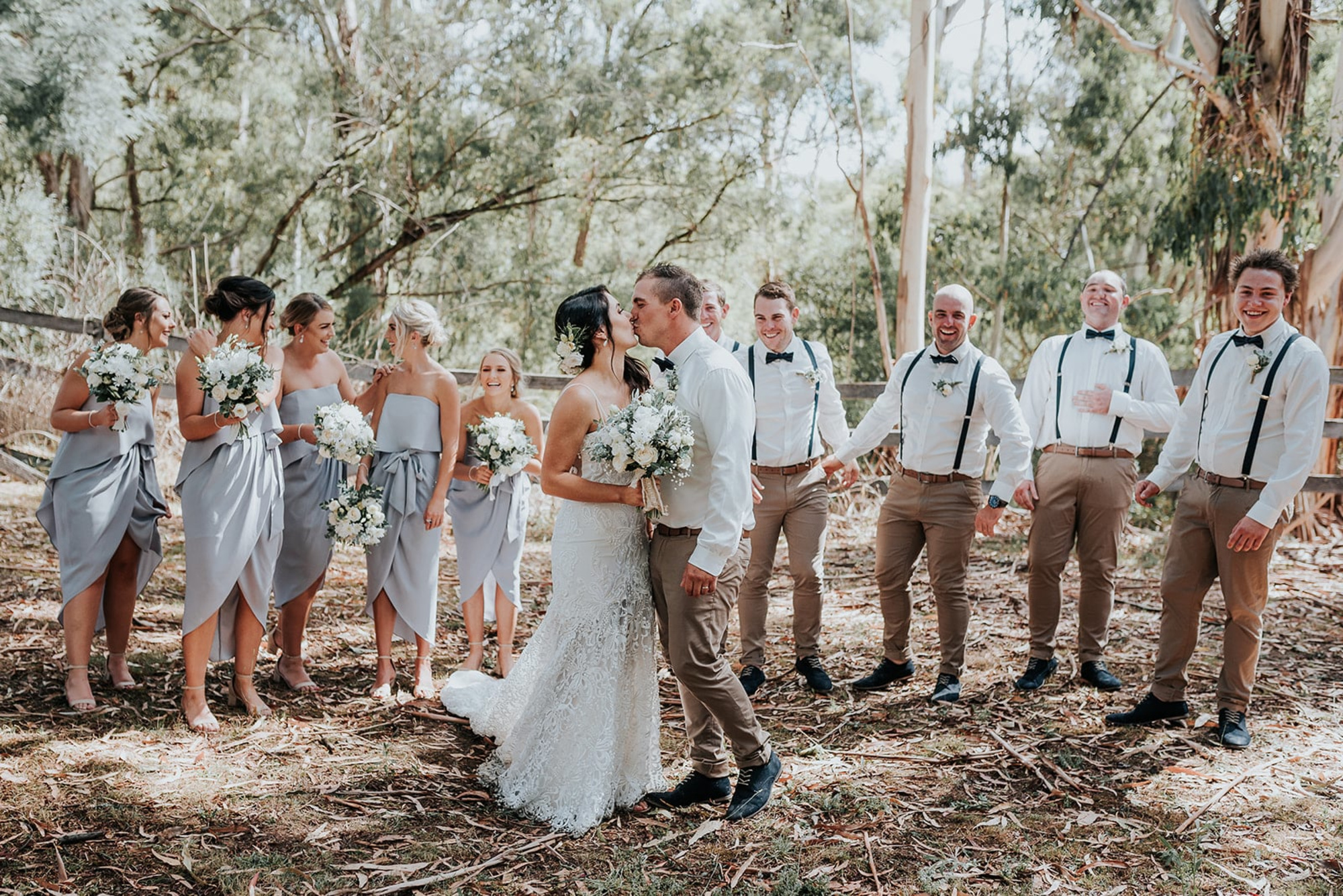 Bride and groom kiss in a woodland setting surrounded by smiling bridesmaids and groomsmen.