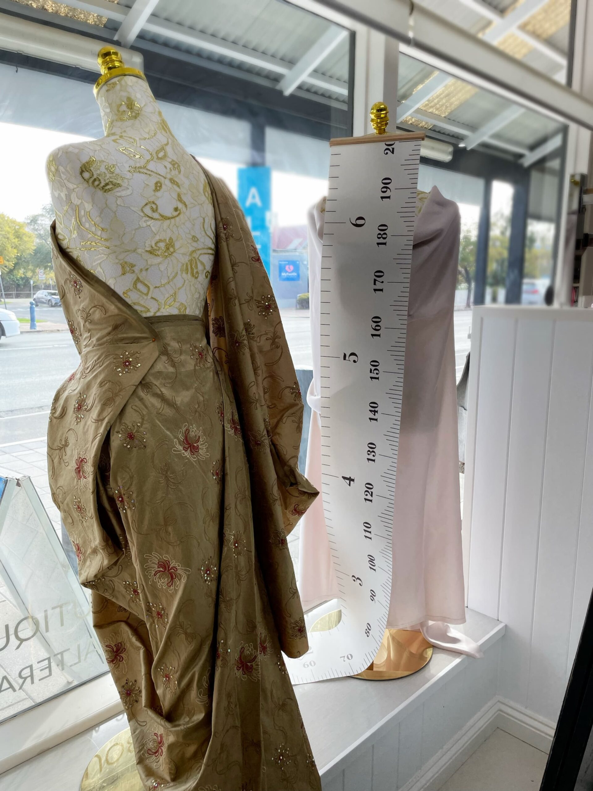 Mannequins in a bridal boutique window displaying a gold embellished gown beside a tall measuring chart.