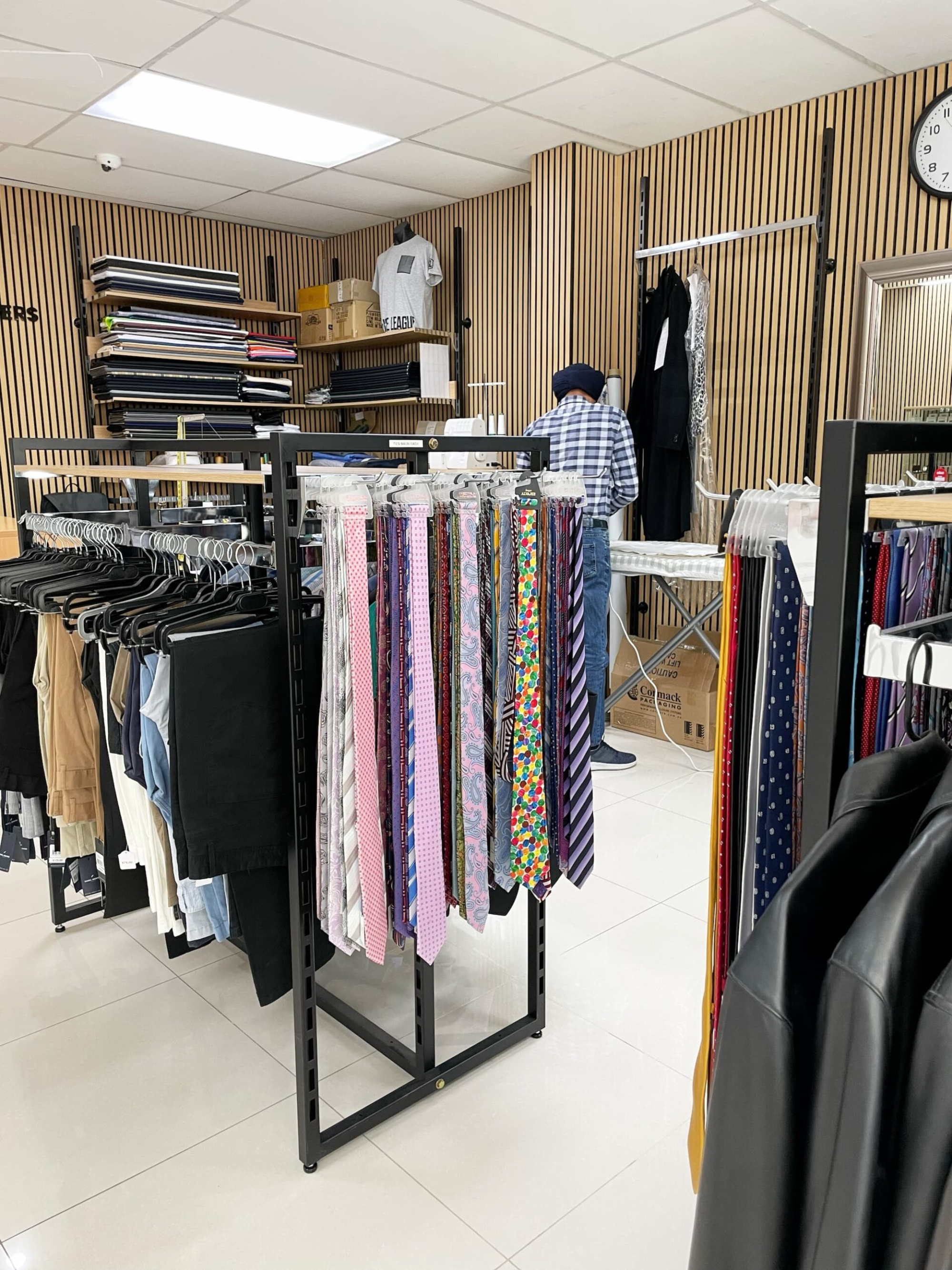 Menswear shop interior with racks of colorful neckties, clothing, and a tailor working in the background.