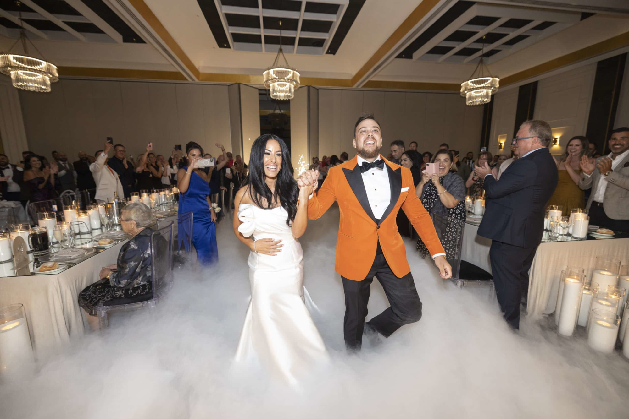 Newlywed couple makes a joyful grand entrance through low-lying fog at a candlelit indoor wedding reception.