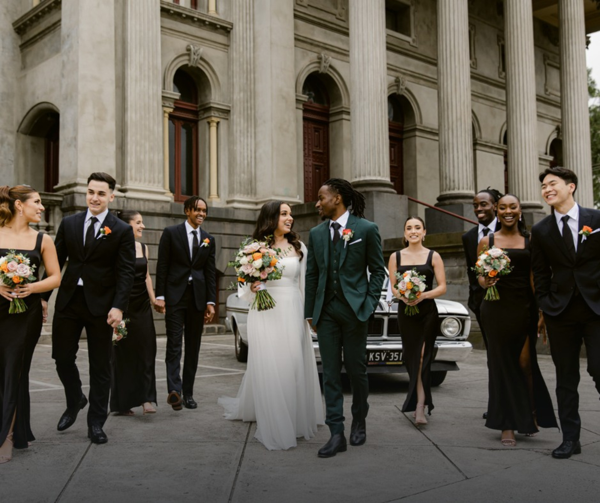 Bride and groom walk with their wedding party holding bouquets in front of a grand stone building.