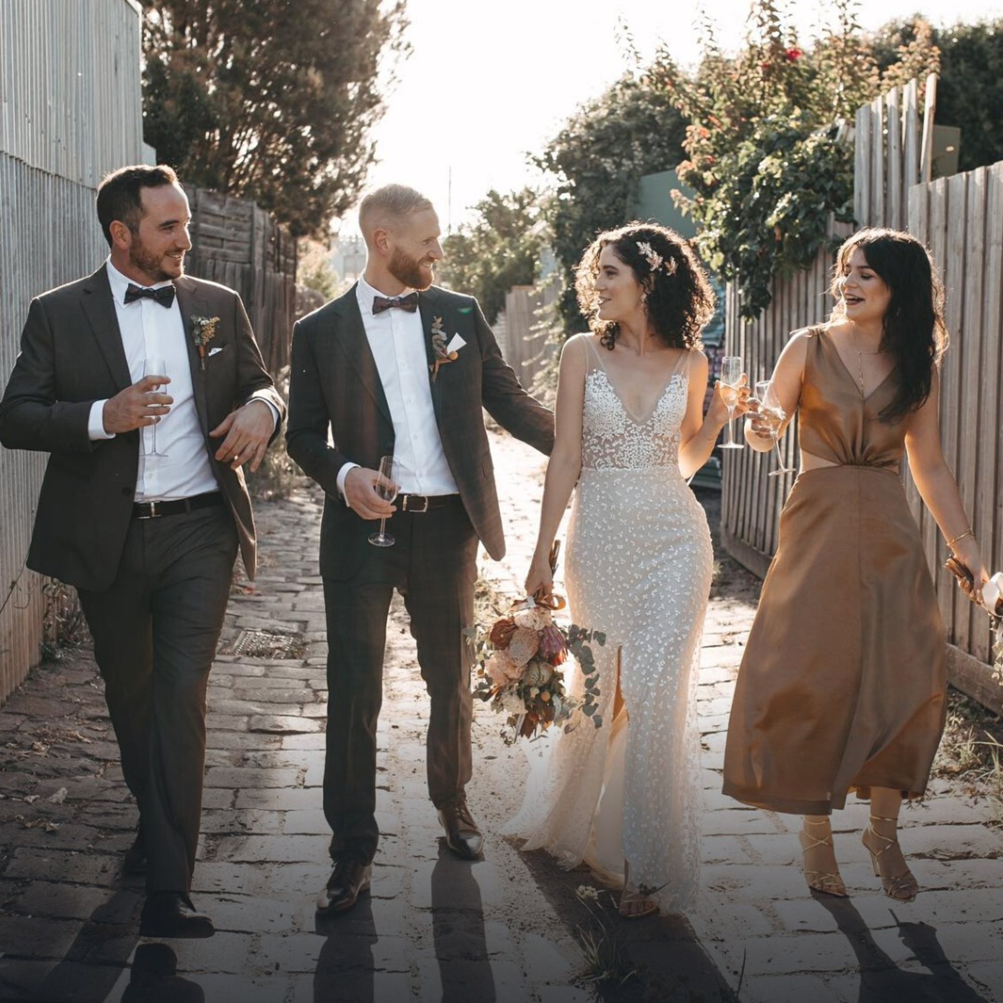 Bride and groom walk down a rustic path with their wedding party, laughing and holding champagne at sunset.