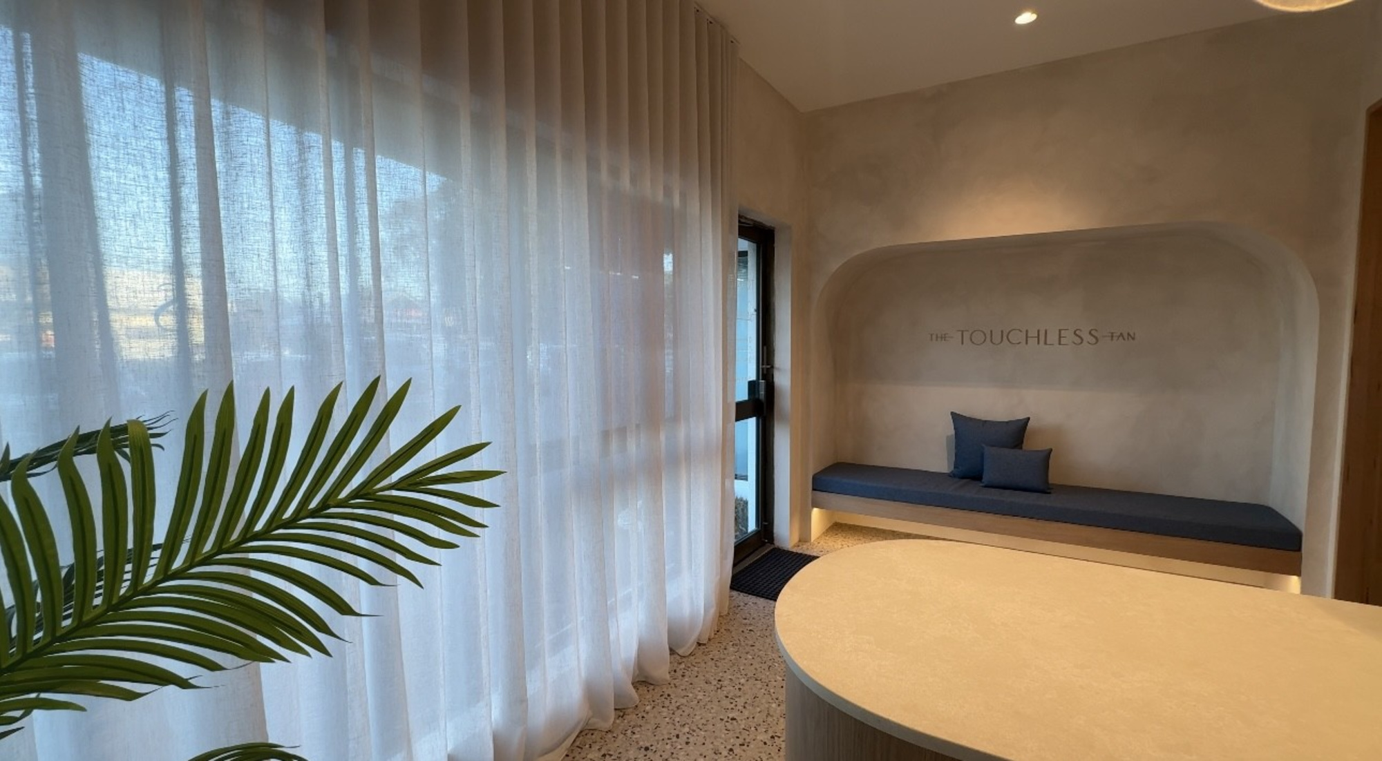 Minimalist salon lounge with sheer curtains, a blue cushioned bench, and The Touchless Tan sign on the wall.