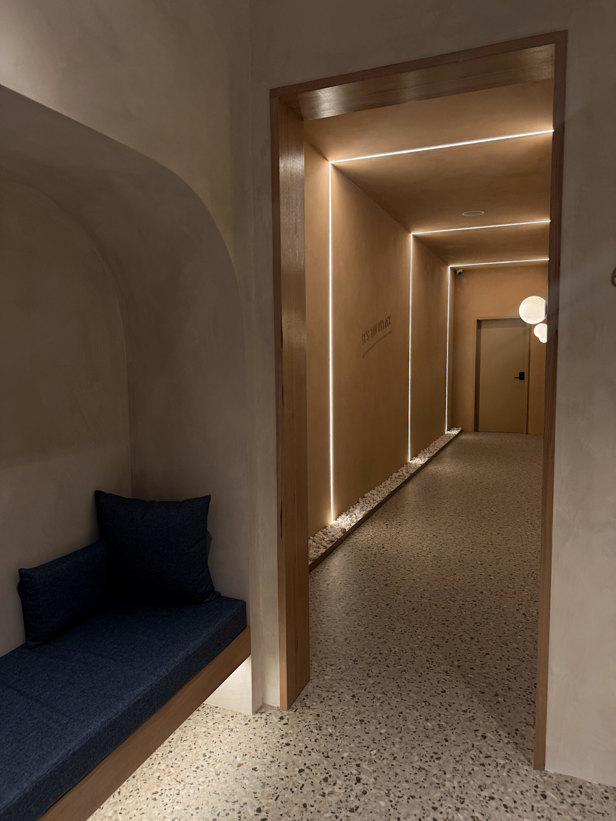 Minimalist hallway with warm lighting, terrazzo floor, and a built-in bench with dark cushions.