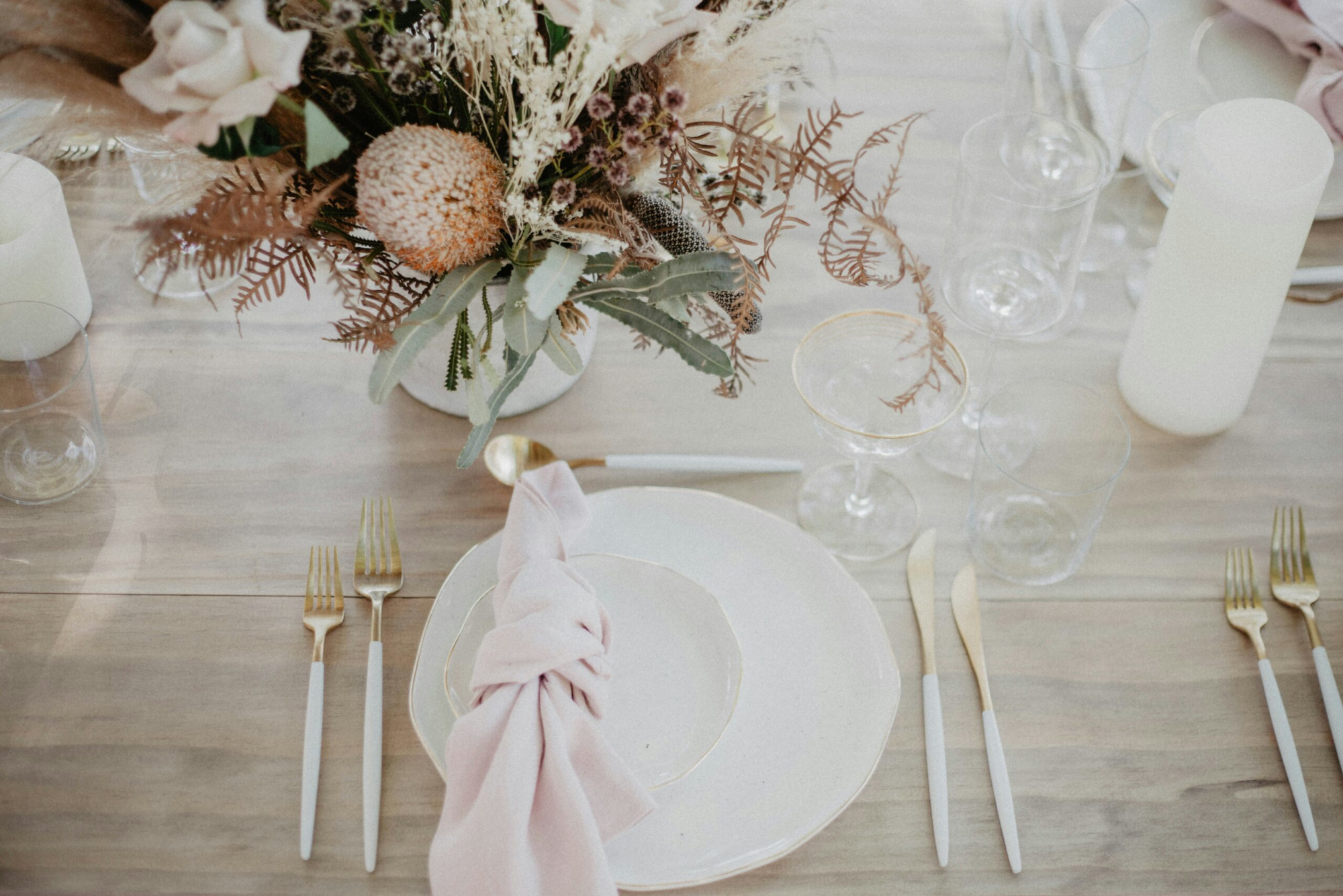 Soft neutral wedding table setting with dried floral centerpiece, gold flatware, and blush knotted napkin.