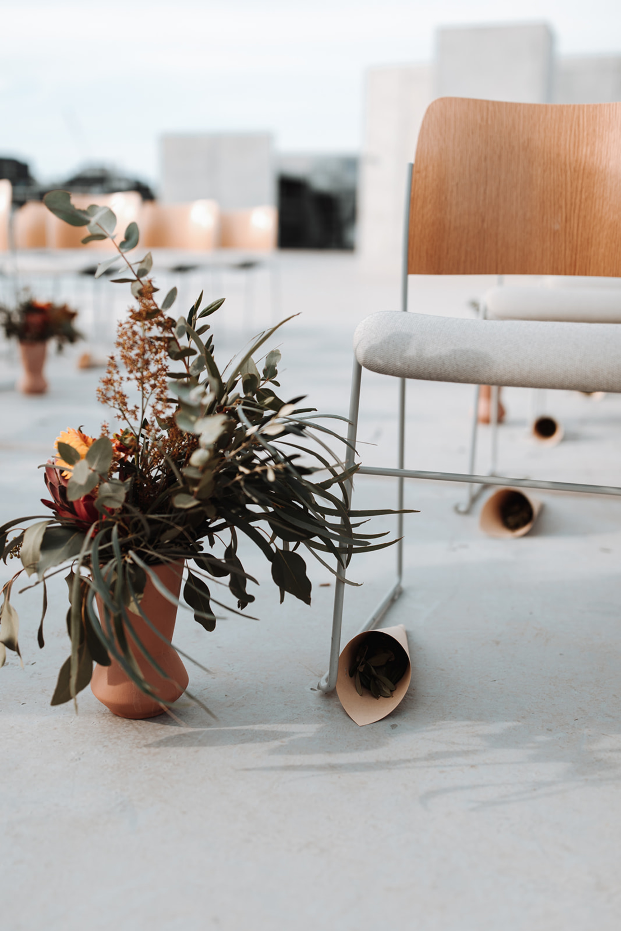 Modern wedding ceremony chairs with terracotta vases of greenery and scattered paper cones on a minimal concrete floor.