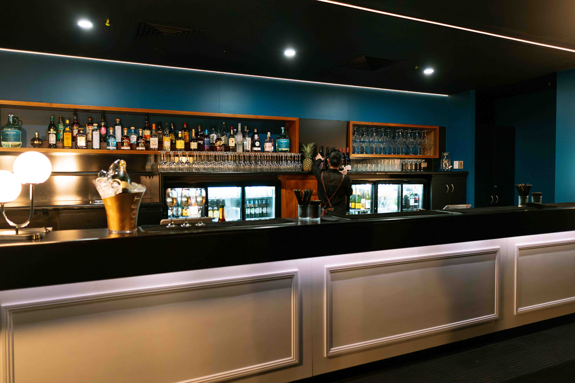 Stylish indoor wedding bar with backlit shelves of liquor, glassware, and a bartender preparing drinks.