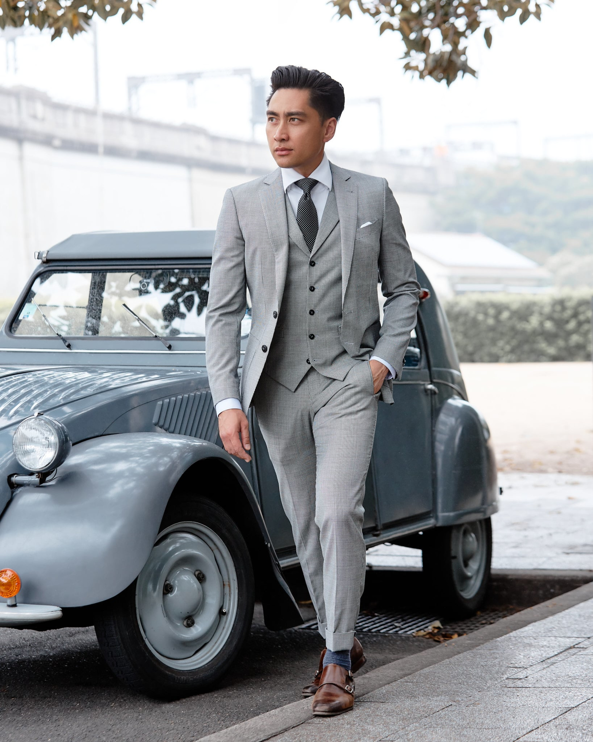 Groom in a tailored light grey three-piece suit poses beside a vintage car in an urban outdoor setting.