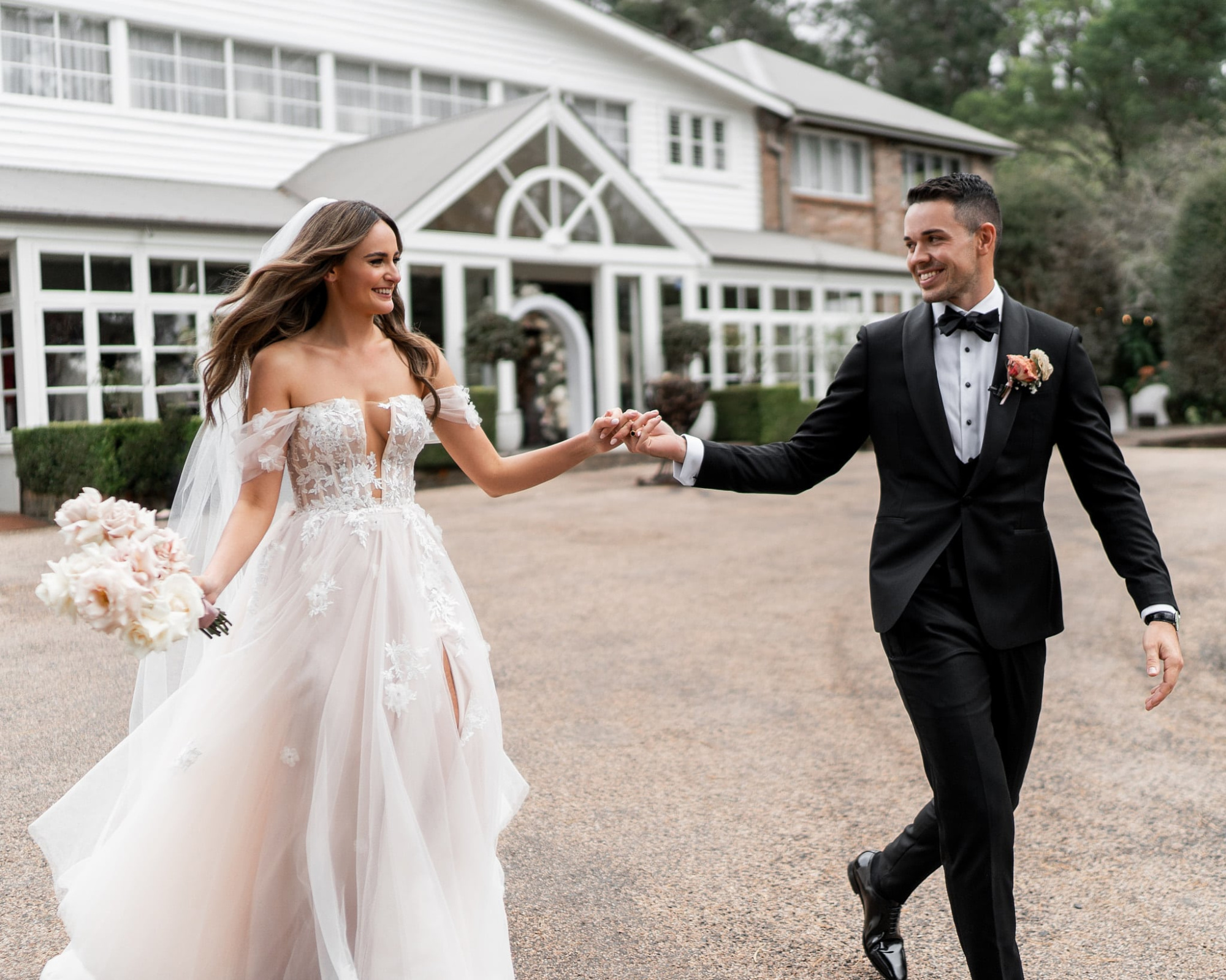 Bride and groom smiling and holding hands while walking outside an elegant white wedding venue.