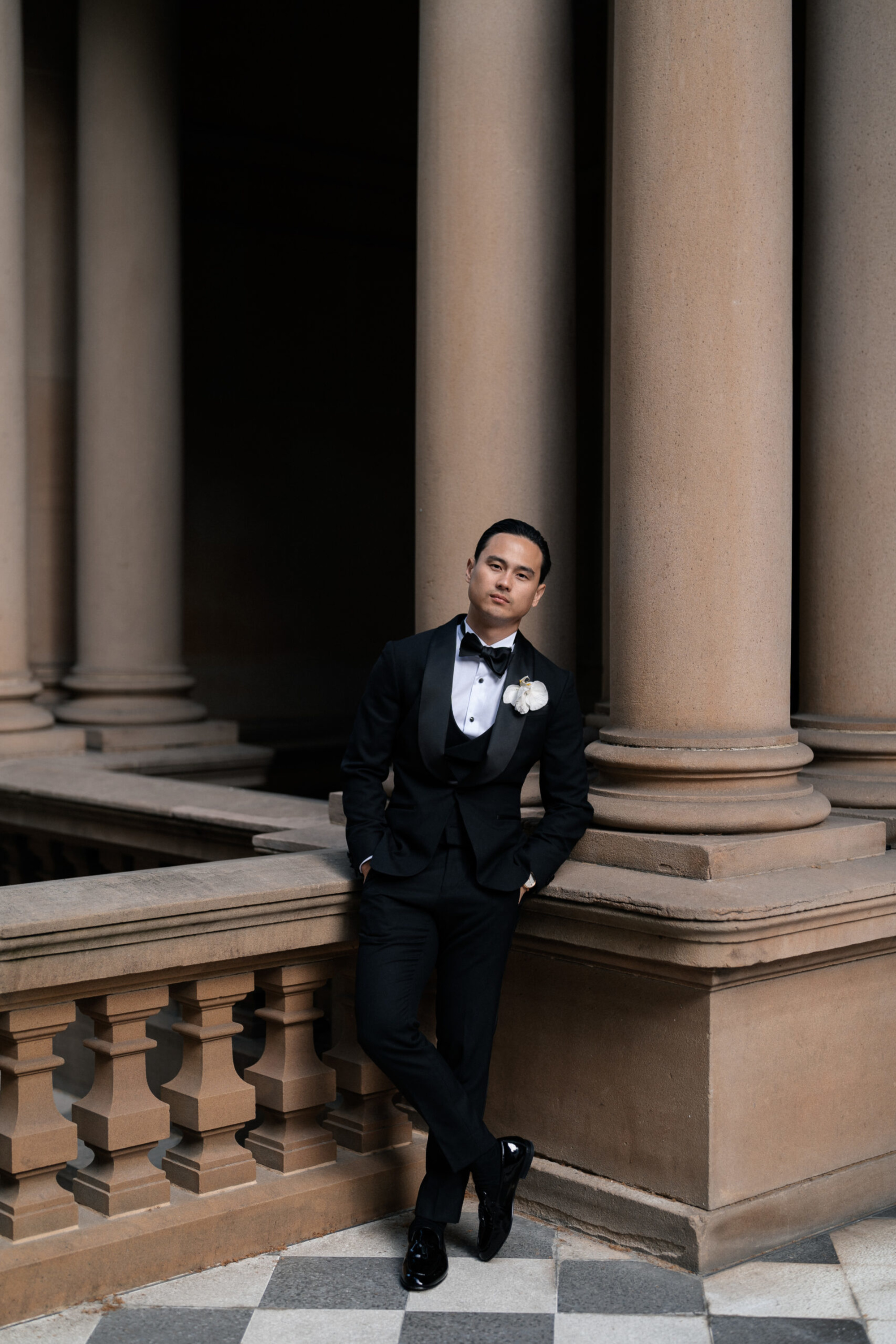 Groom in a black tuxedo leans against stone columns at an elegant architectural venue.
