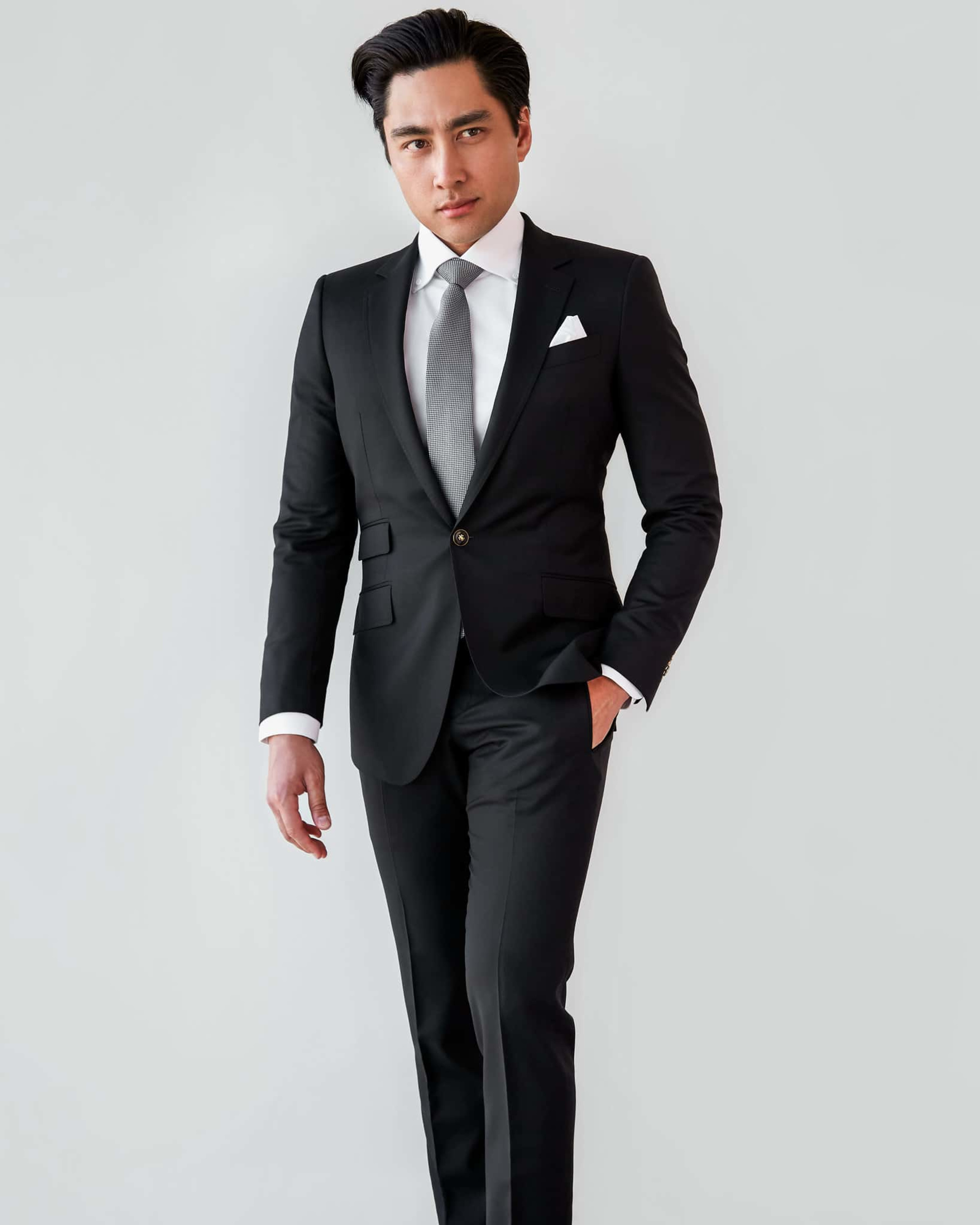 Well-dressed man in a tailored black suit and tie against a light background.