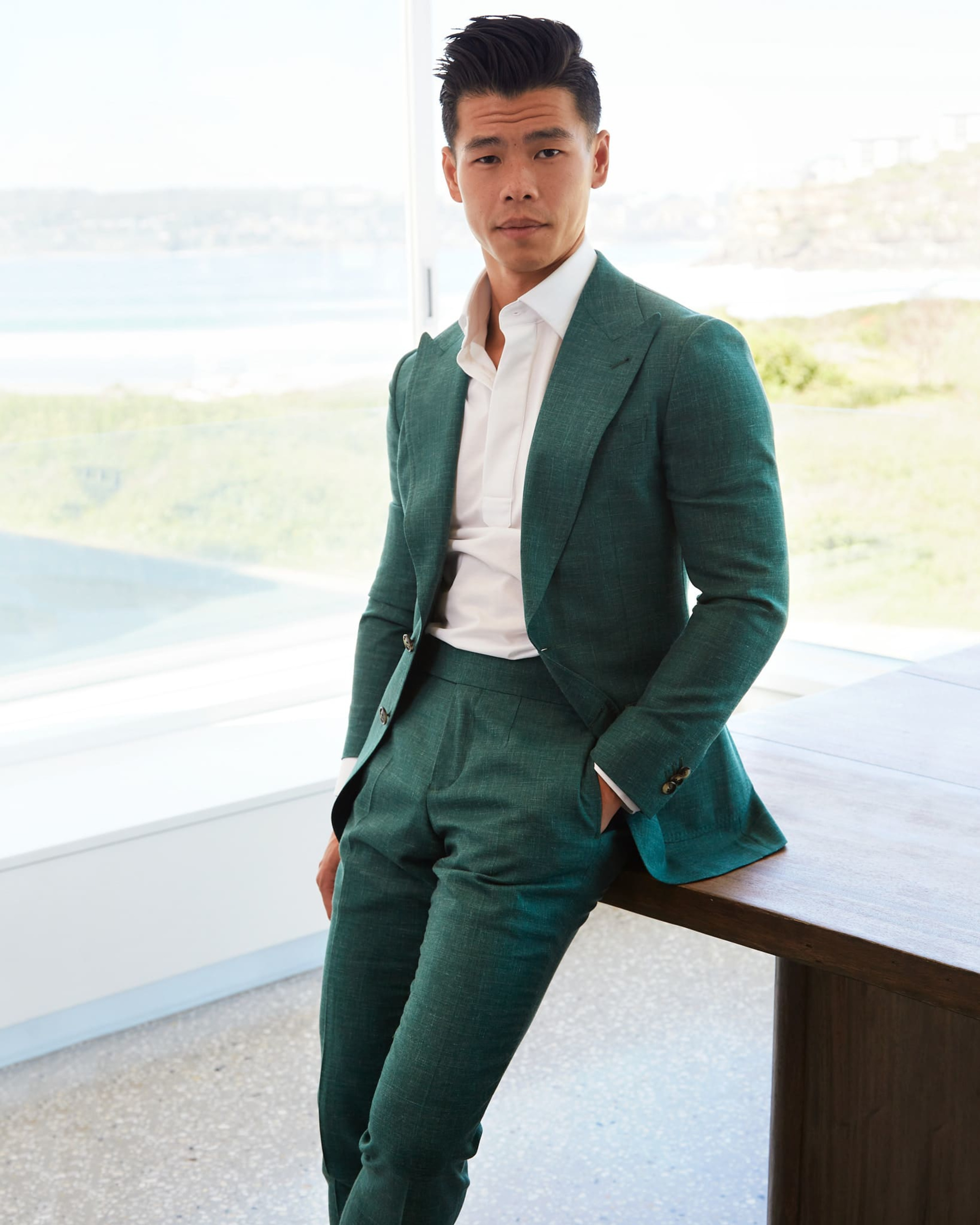Man in a tailored green suit and white shirt poses indoors with a bright coastal view behind him.