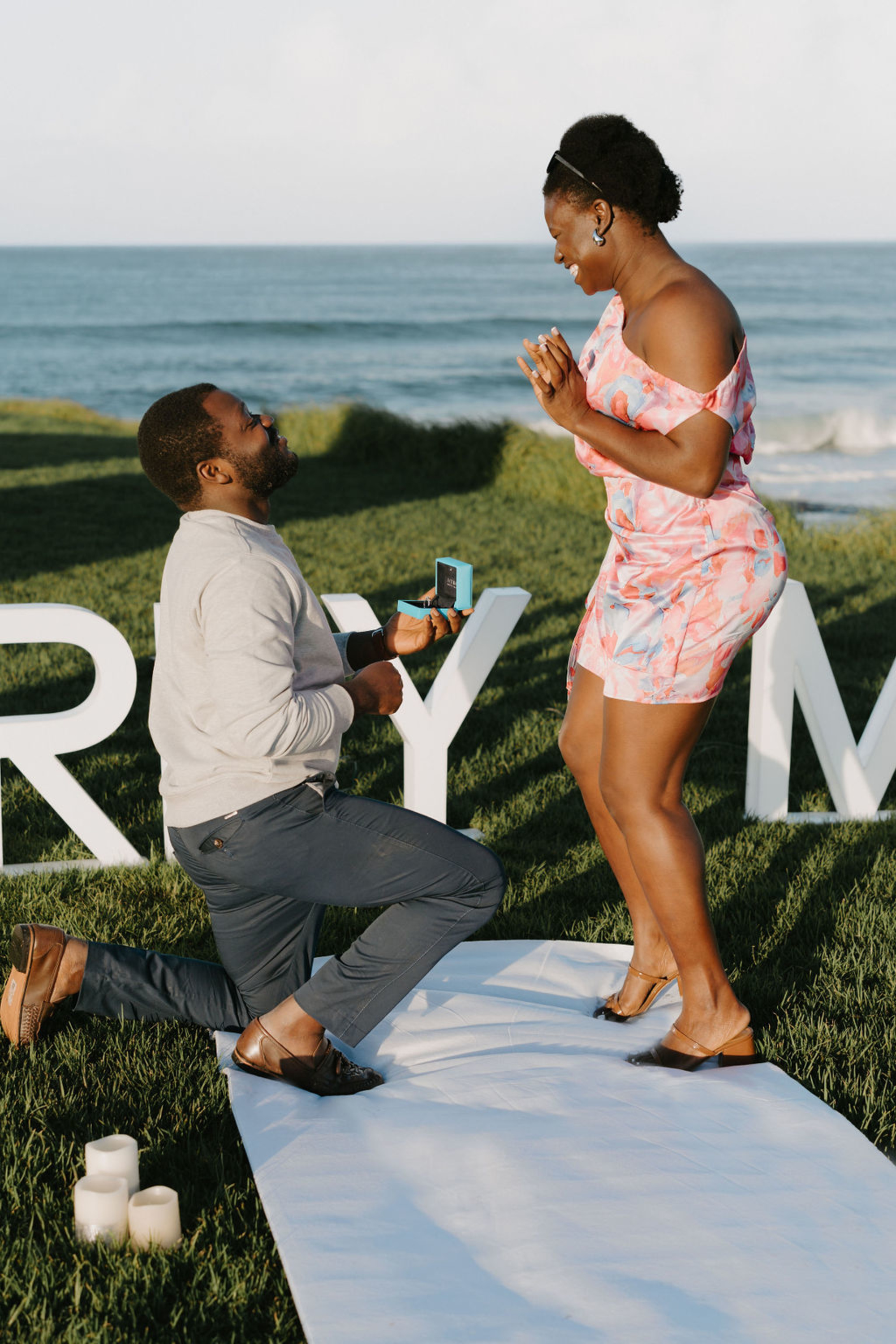 Man proposes on one knee to his joyful partner during a beachside engagement setup at sunset.