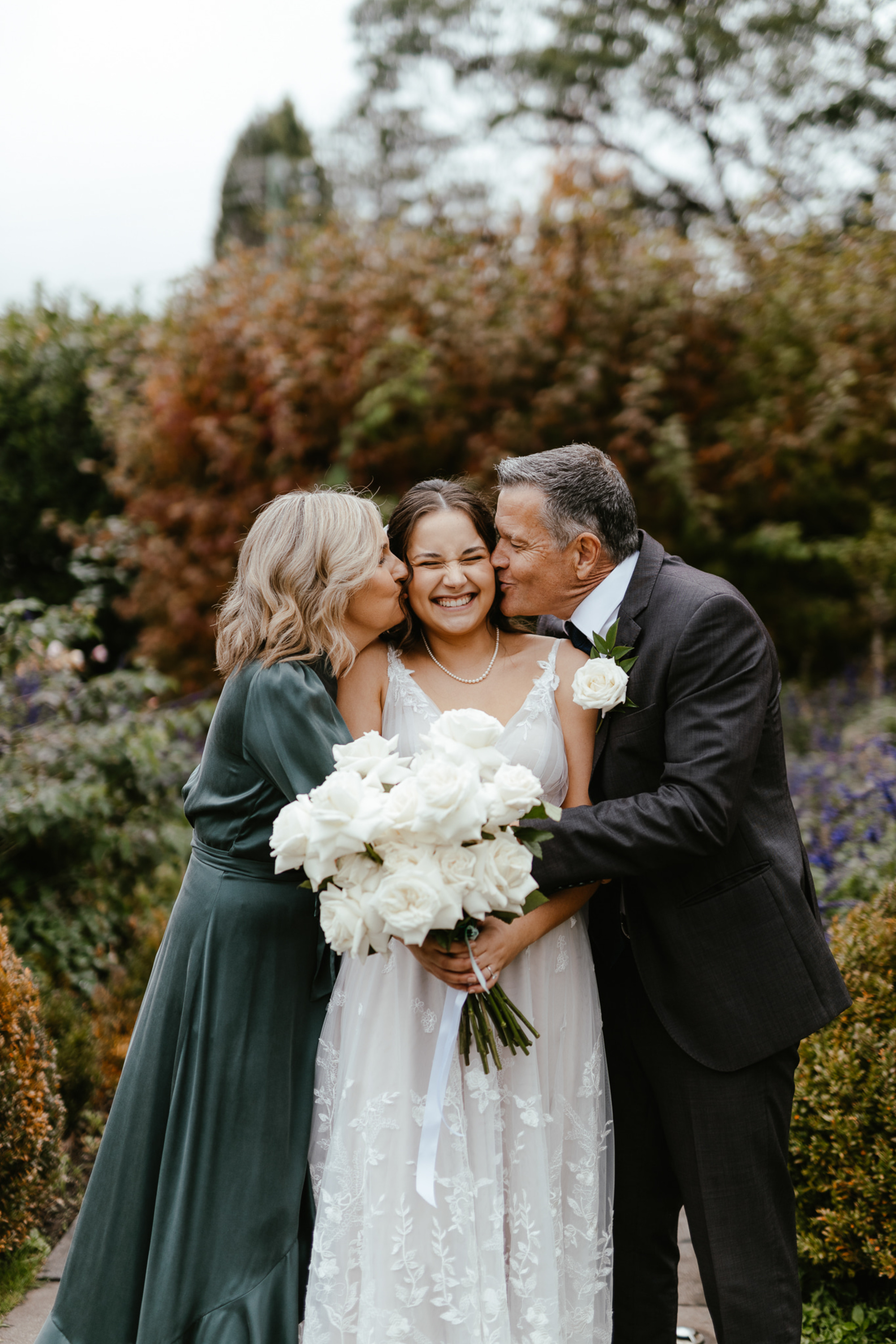 Bride holding white rose bouquet as her parents kiss her cheeks in a lush garden setting.