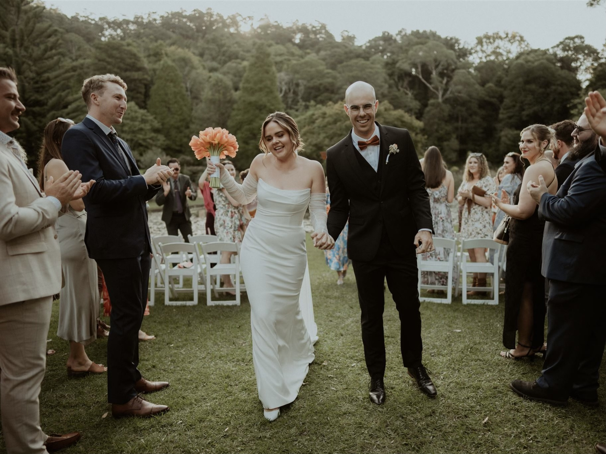 Newlywed couple walks joyfully down an outdoor aisle as guests applaud in a lush garden setting.