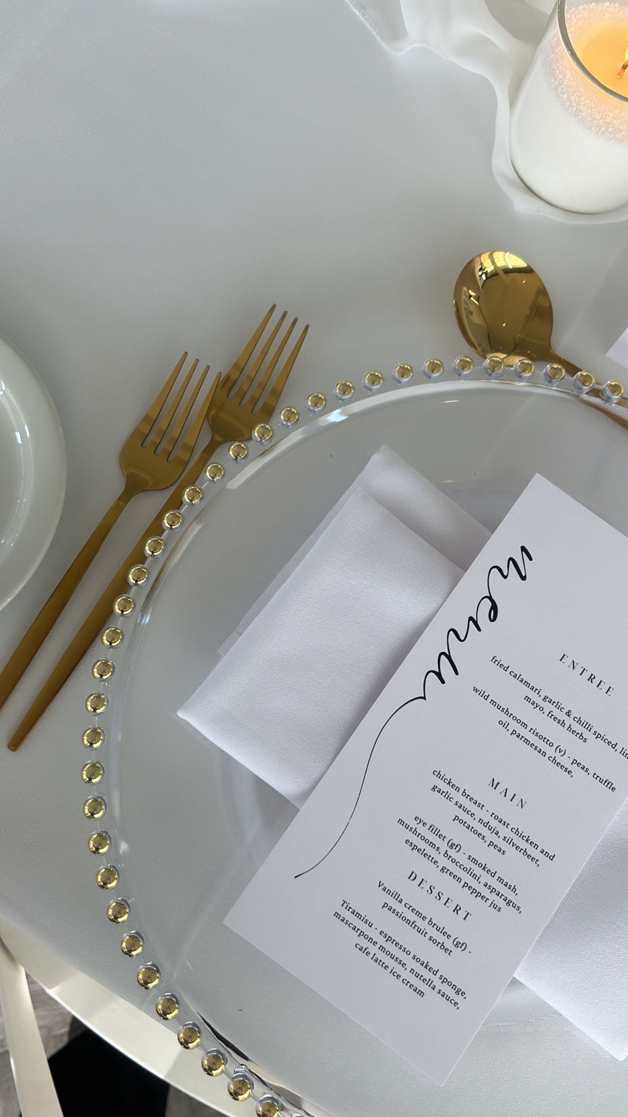 Elegant wedding place setting with gold cutlery, jeweled charger plate, white napkin, printed menu, and candle on a white tablecloth.