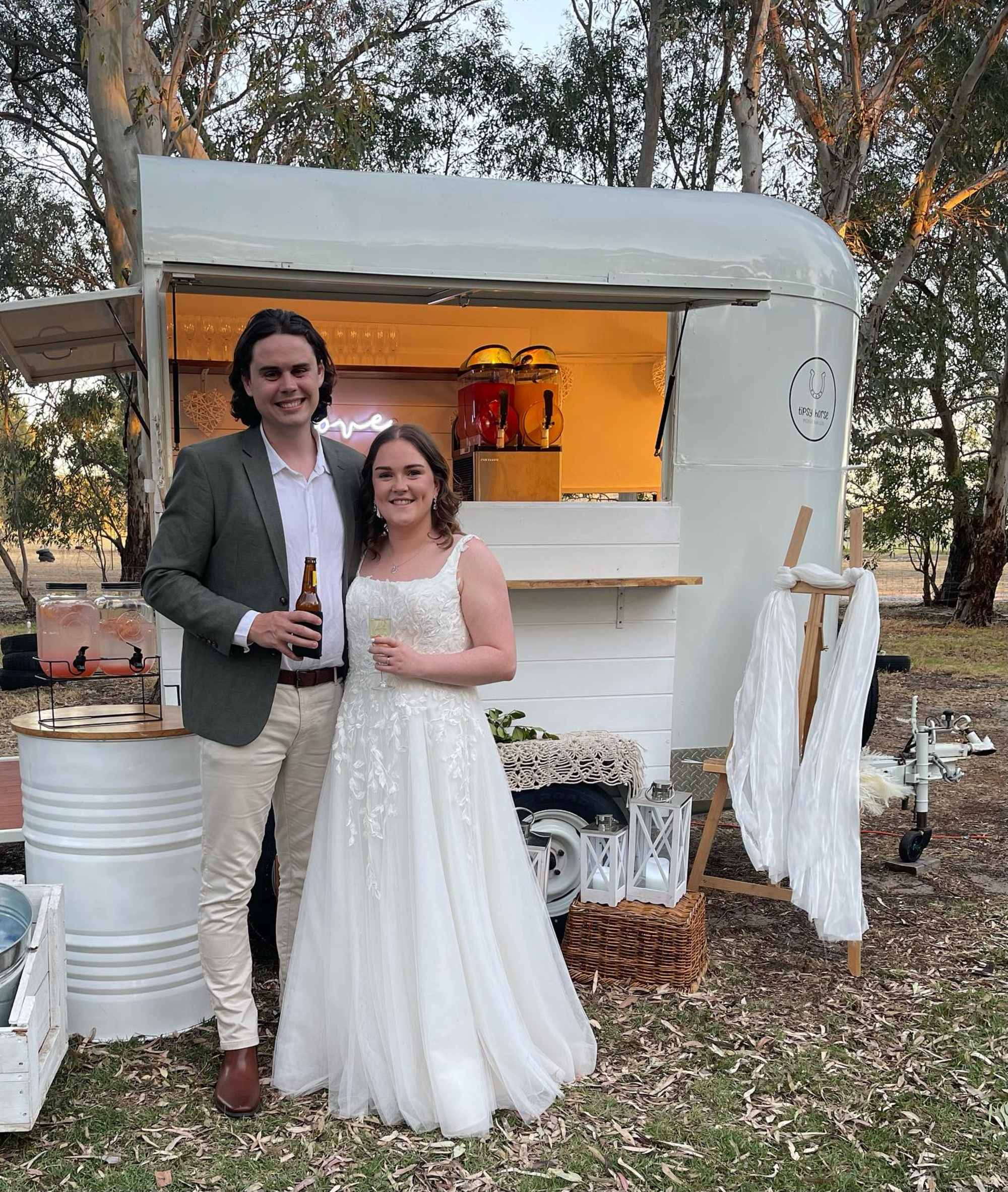 Bride and groom stand smiling in front of a rustic mobile bar trailer at an outdoor wedding reception.