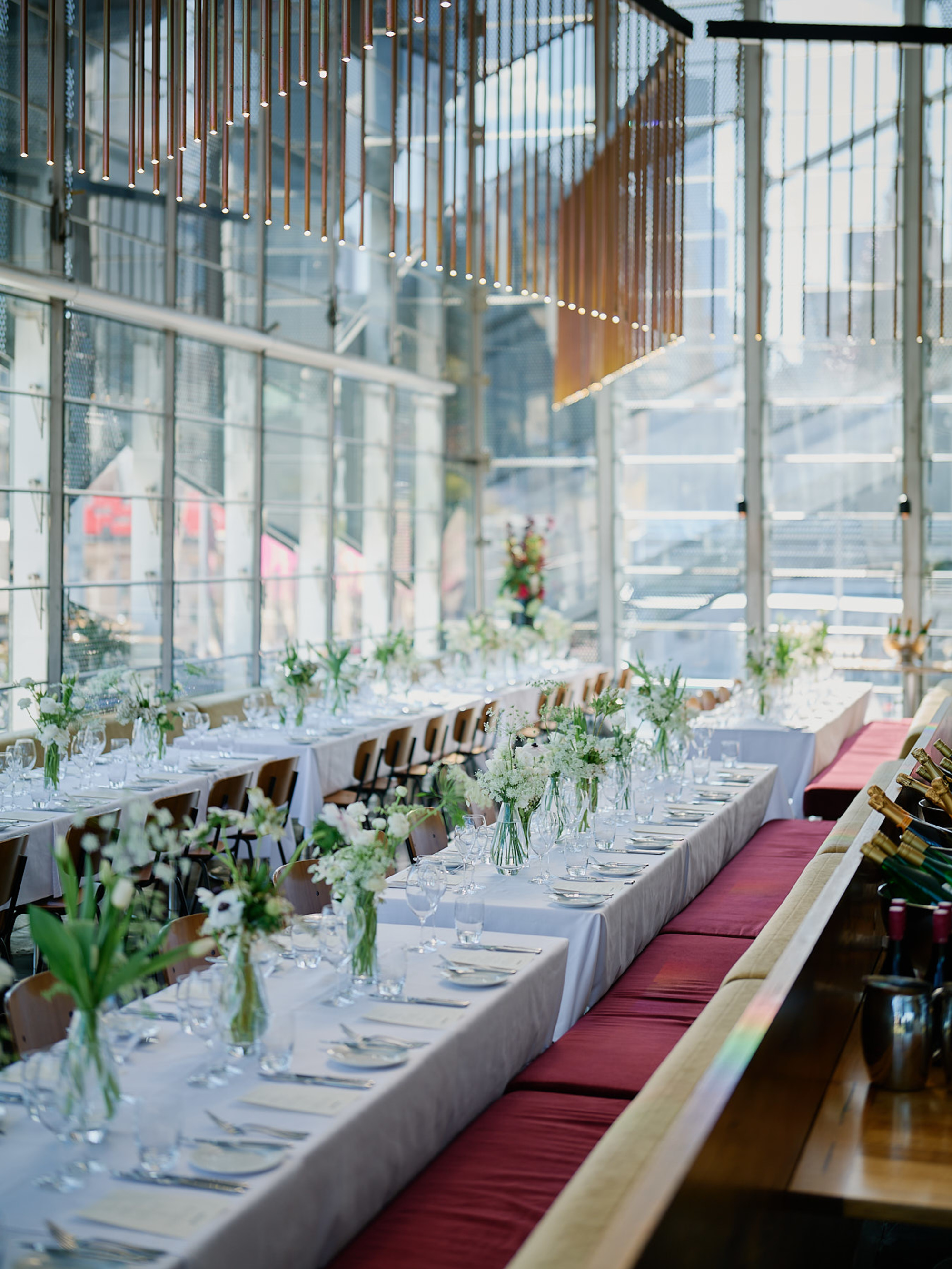 Modern glass-walled wedding reception space with long banquet tables, white linens, and green and white floral centerpieces.