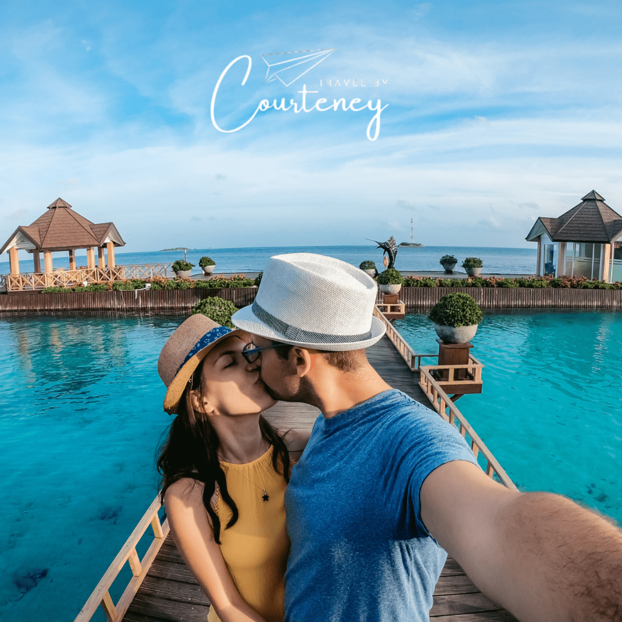 Couple kisses on a tropical overwater boardwalk at a turquoise ocean resort with pavilions in the background.