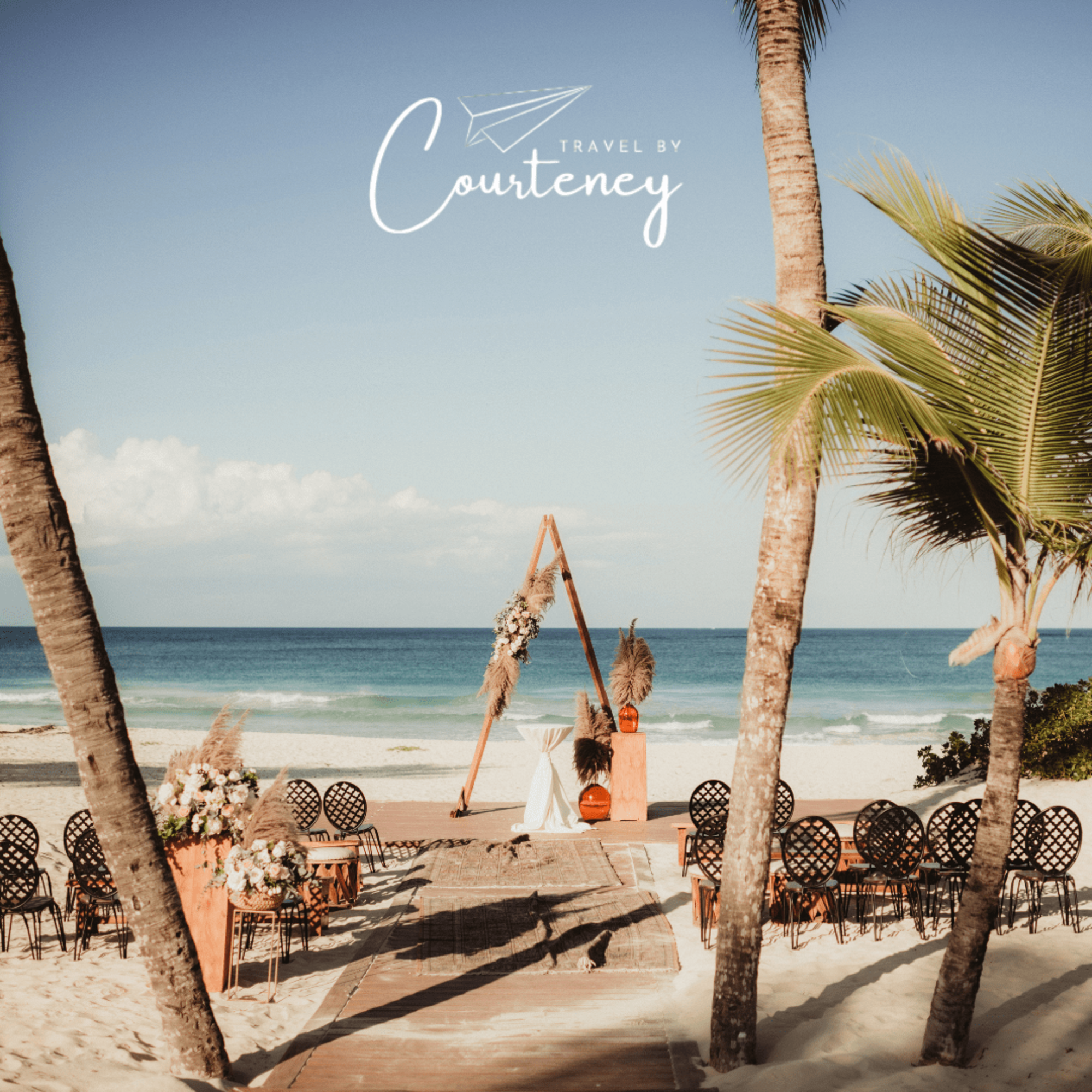 Boho beach wedding ceremony setup with triangle arch and chairs facing the ocean under palm trees.