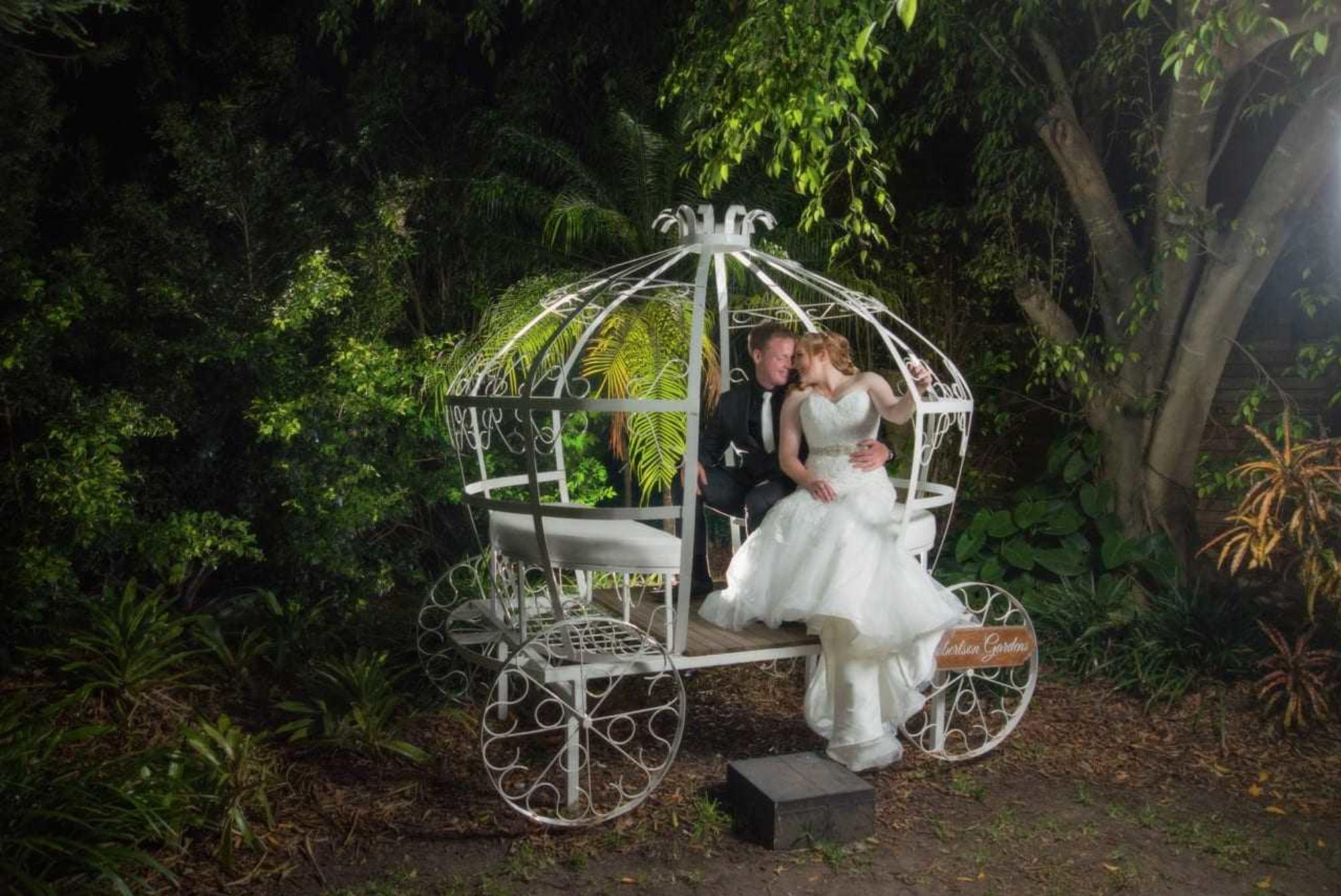 Bride and groom sit in a white fairytale carriage in a lush garden at night.