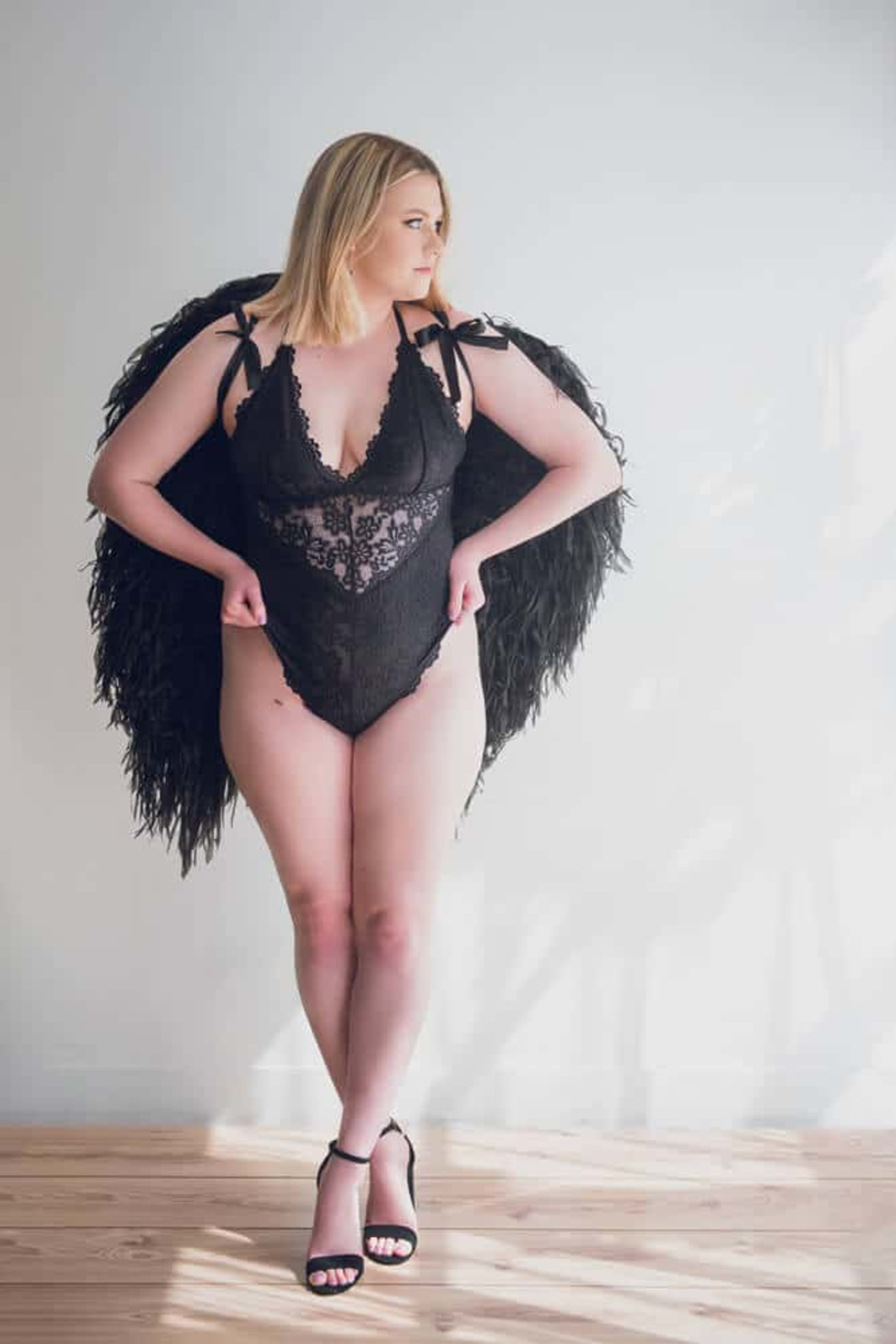 Bride in black lace lingerie and feathered wings poses in bright studio for a boudoir session.