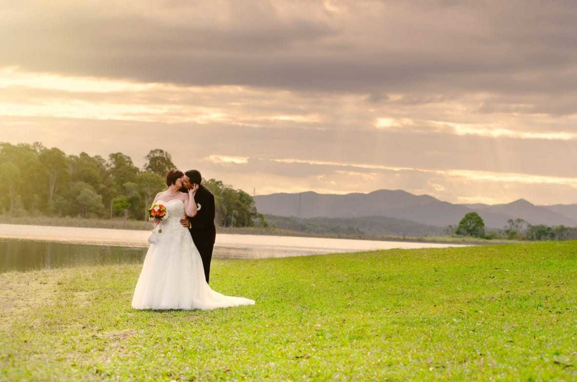 Bride and groom kiss by a lakeside meadow at sunset with mountains and trees in the background.