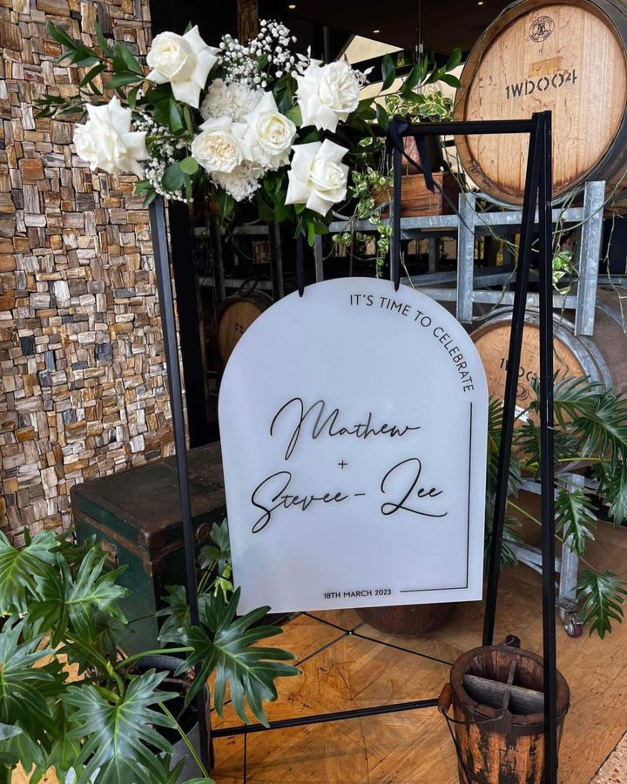 Modern wedding welcome sign with white roses and greenery displayed in front of wooden wine barrels.