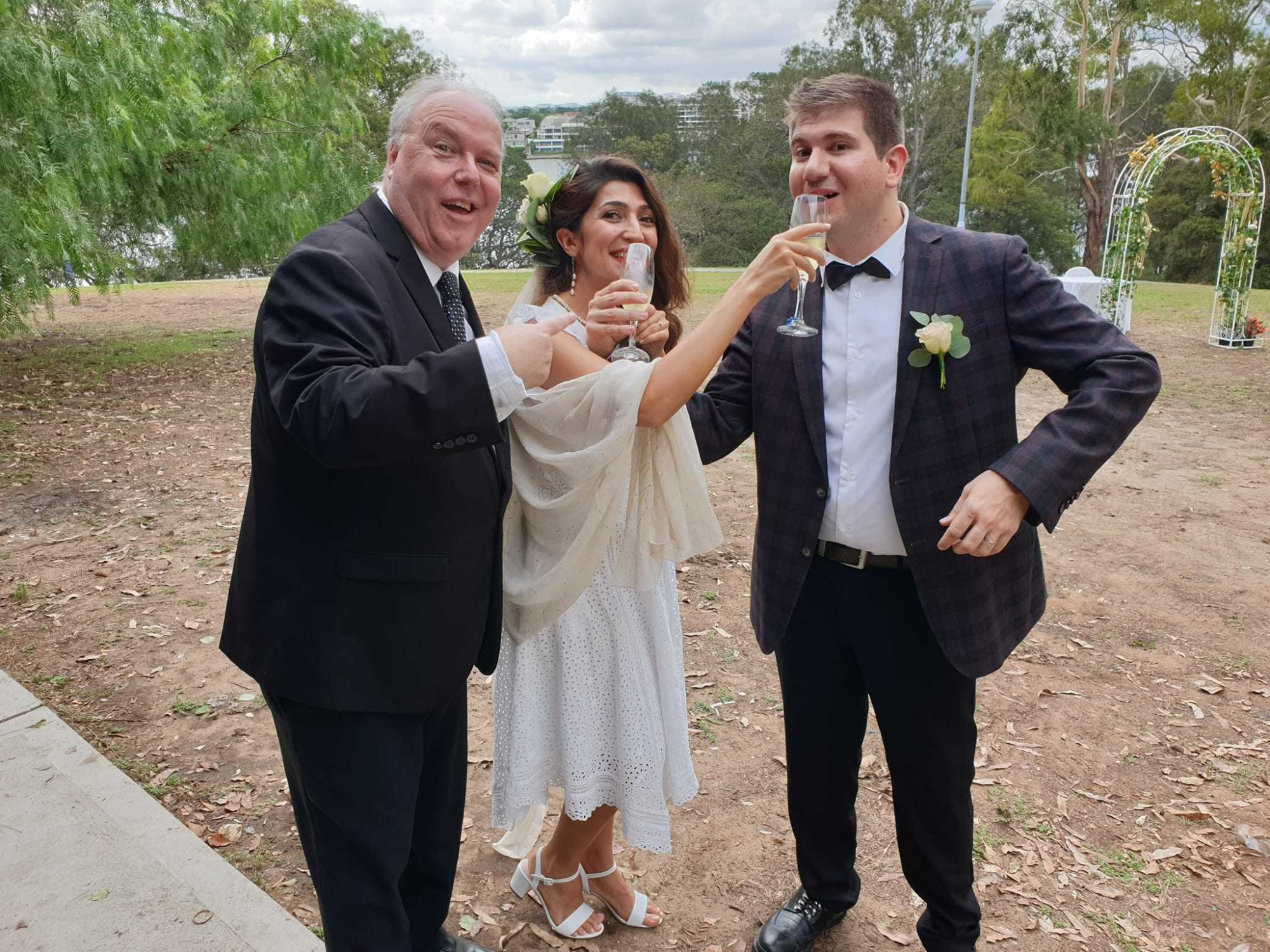 Bride and groom share a champagne toast with a guest at an outdoor park wedding ceremony.
