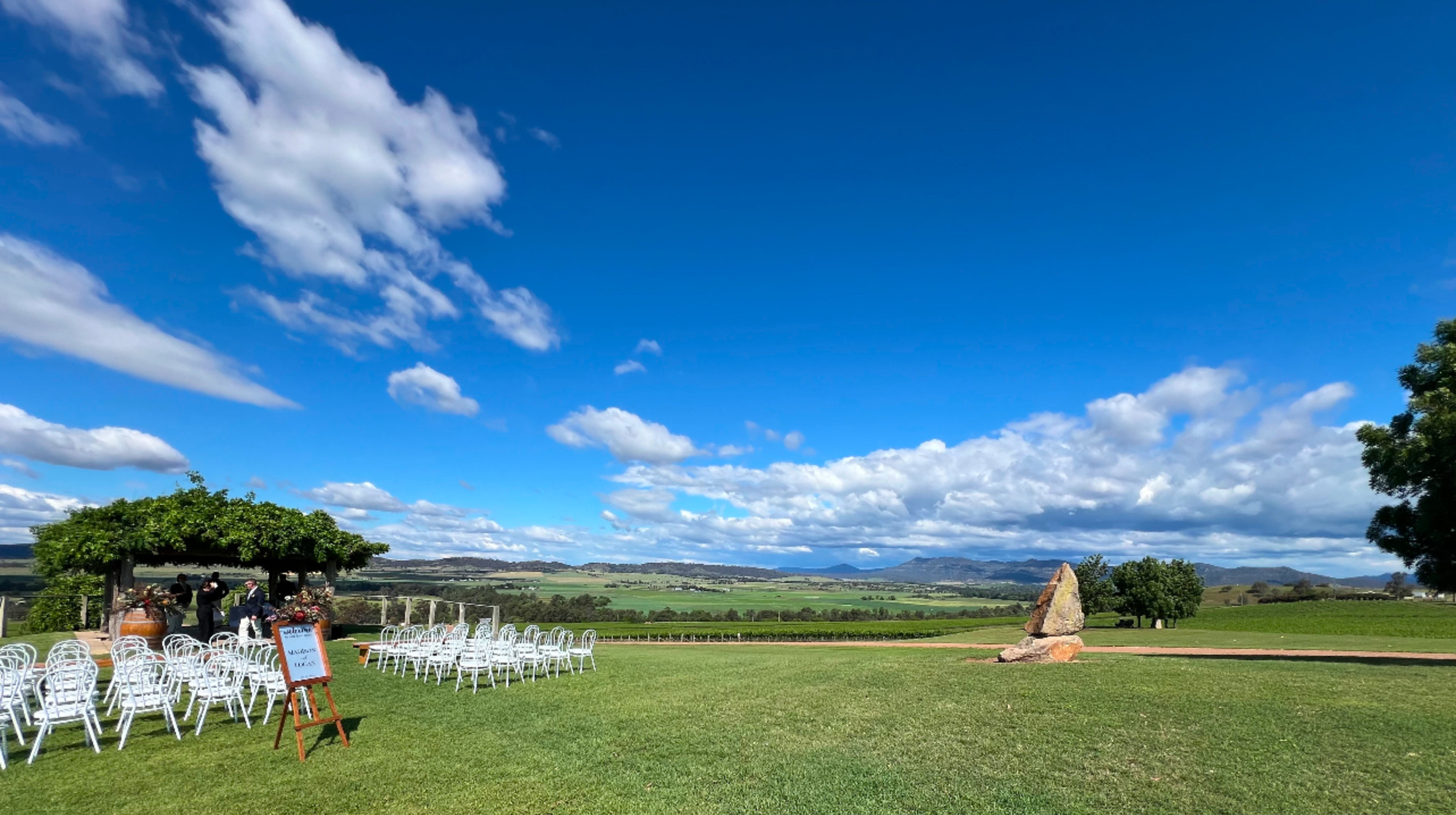 Outdoor wedding ceremony setup on a lush lawn overlooking rolling countryside and mountains under a bright blue sky.