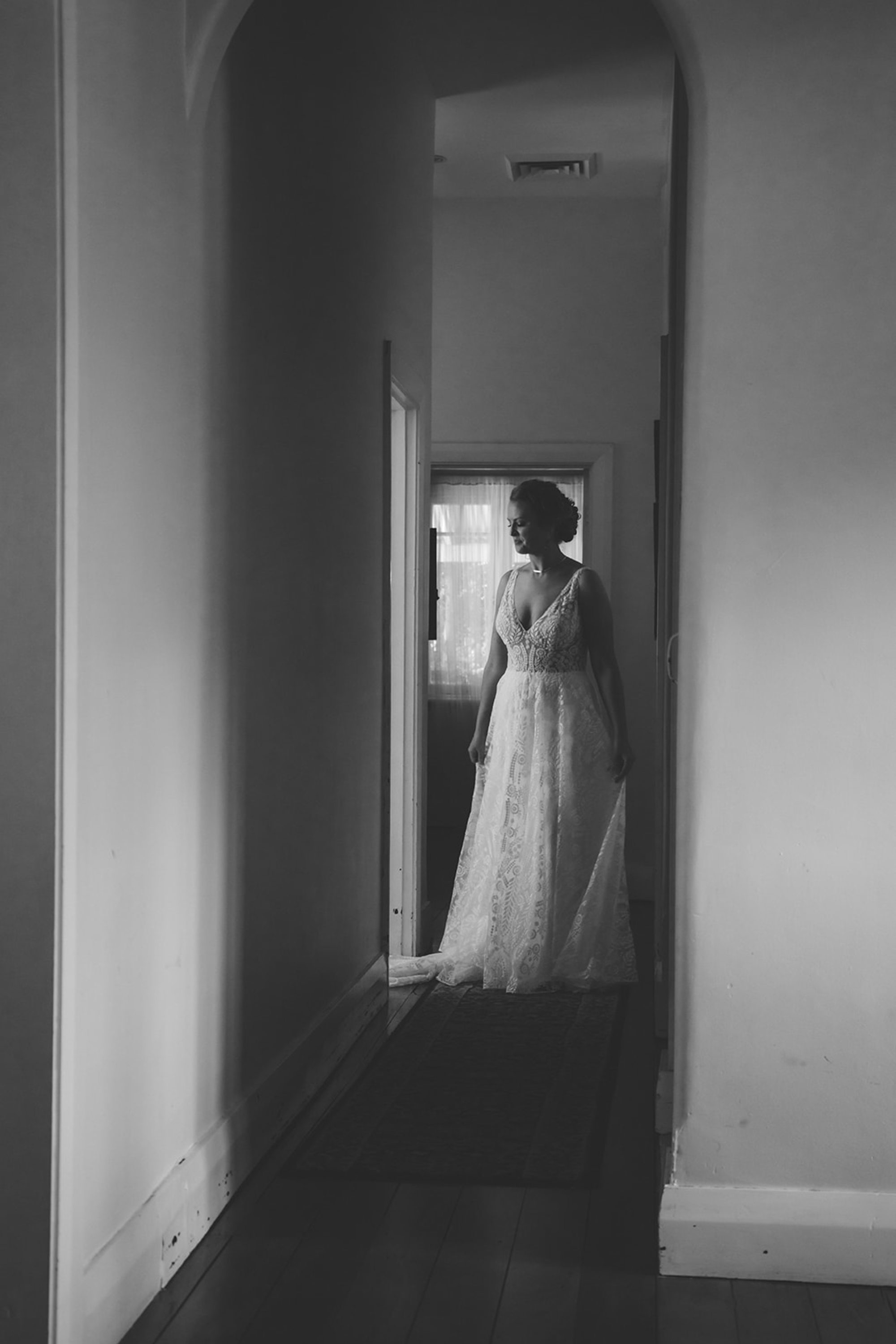 Black and white photo of a bride in a lace gown standing thoughtfully in a hallway by a softly lit doorway.