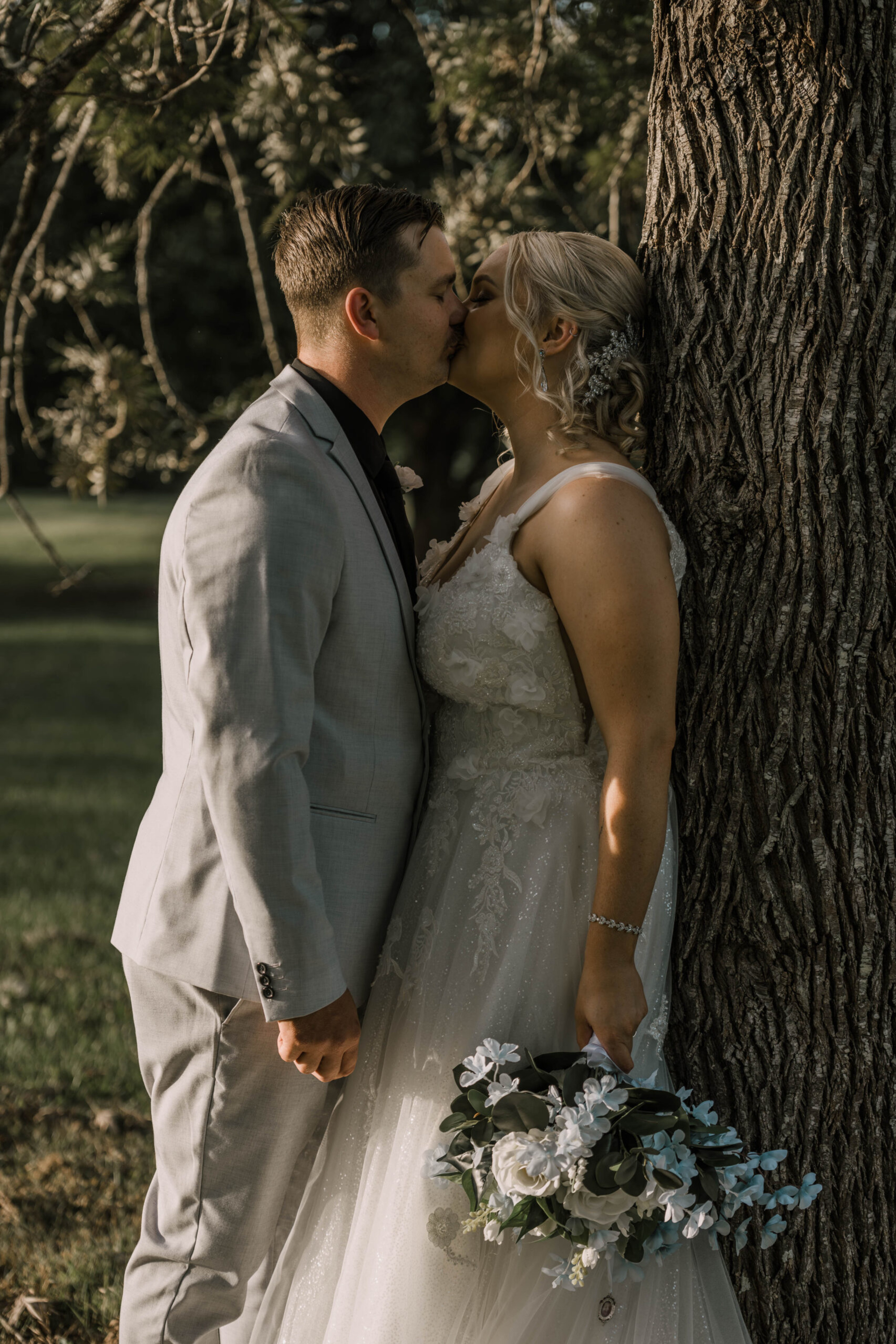 Bride and groom share a kiss by a tree, holding a white and green bouquet in soft outdoor light.