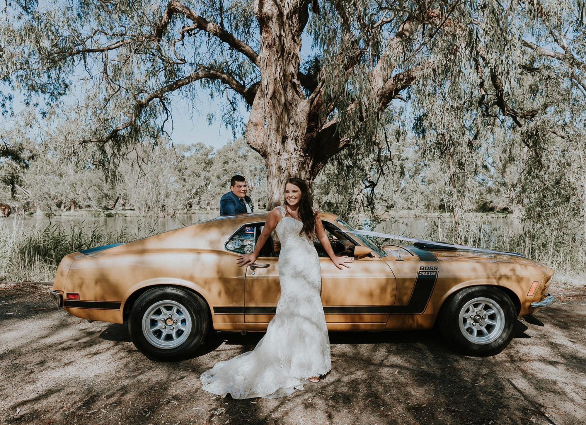 Bride in lace gown posing with classic car under a large tree while groom stands behind by a lakeside.