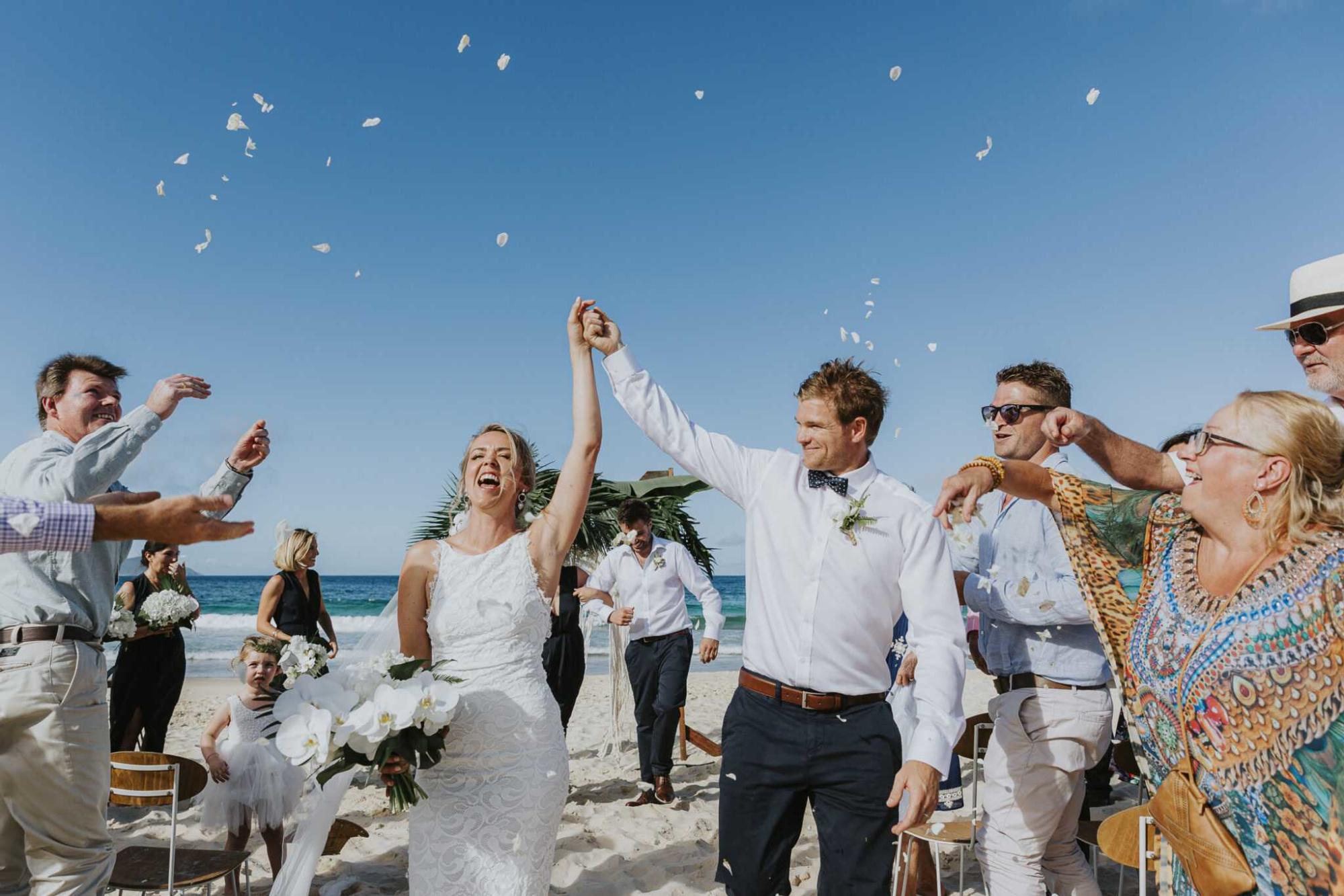 Joyful bride and groom walk down a sandy beach aisle as guests toss flower petals in celebration.