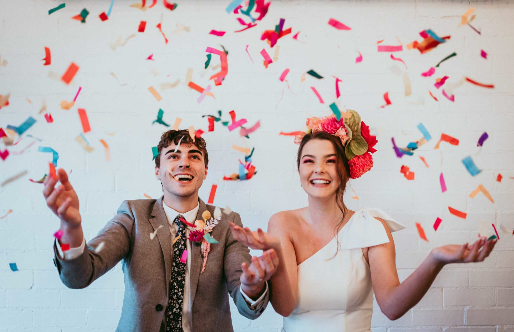 Joyful wedding couple tossing colorful confetti against a white brick wall backdrop.